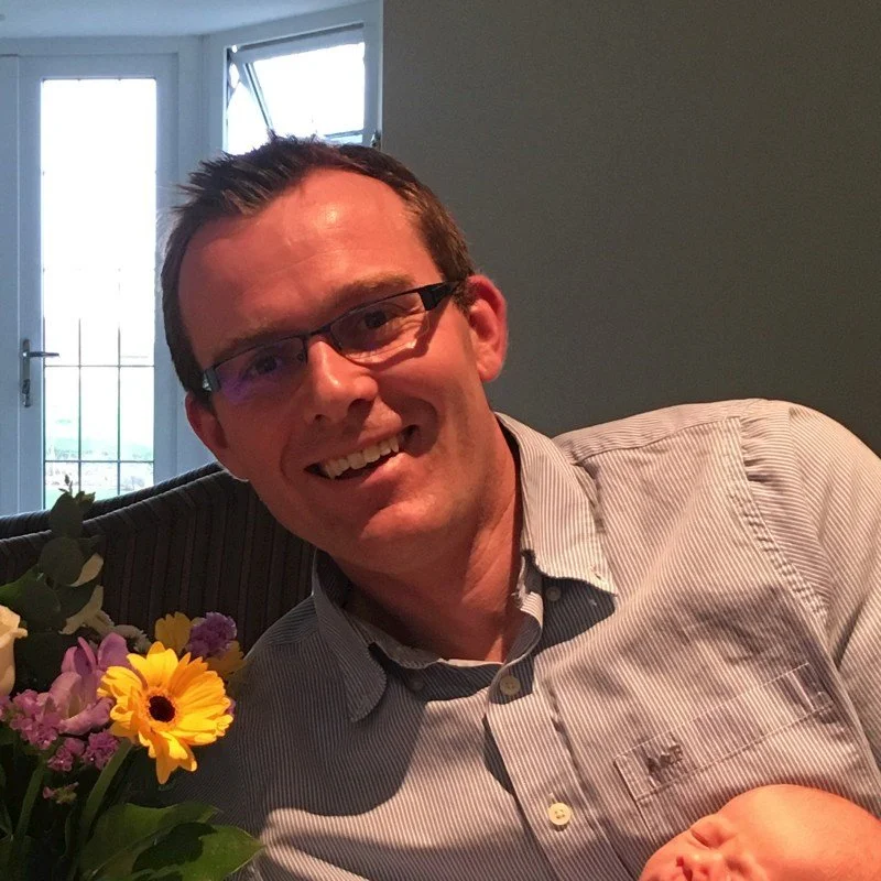 A smiling man wearing glasses and a striped shirt, sitting at a table with a bouquet of colorful flowers, indoors near a window.
