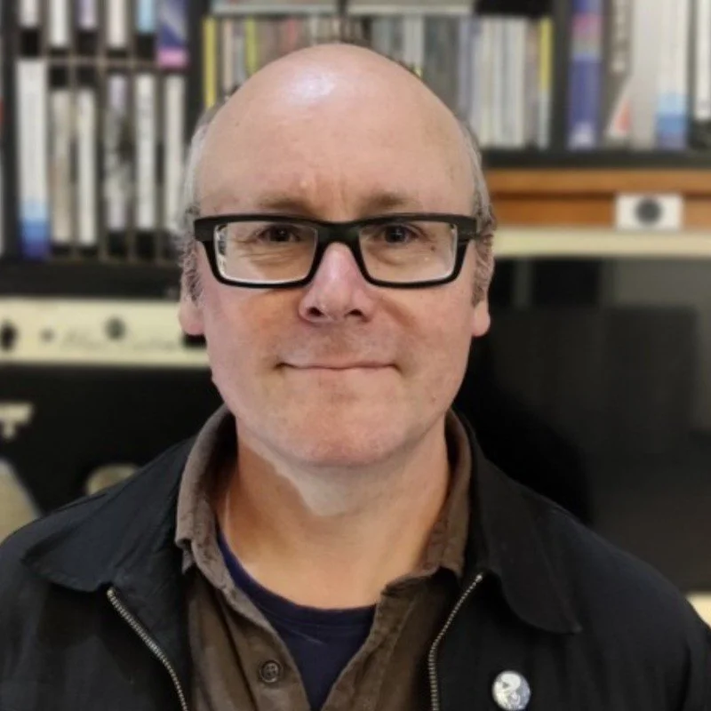 A middle-aged man with glasses, short hair, and a slight smile, standing indoors with a bookshelf and music equipment in the background.