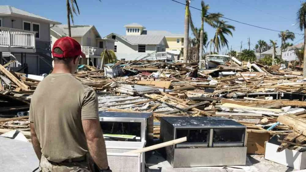 A man wearing a red cap standing amid debris of destroyed buildings after a storm or hurricane, with damaged houses and palm trees in the background.