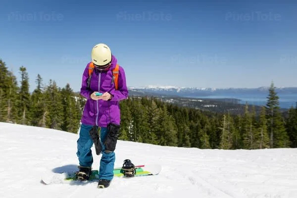 Person in purple jacket, teal pants, and helmet standing on snow with skis, holding a smartphone, in a snowy landscape with trees and mountains in the background.