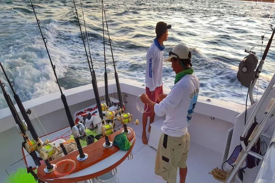 Two men on a boat with fishing rods, one is adjusting fishing gear while the other stands nearby, with water visible in the background.