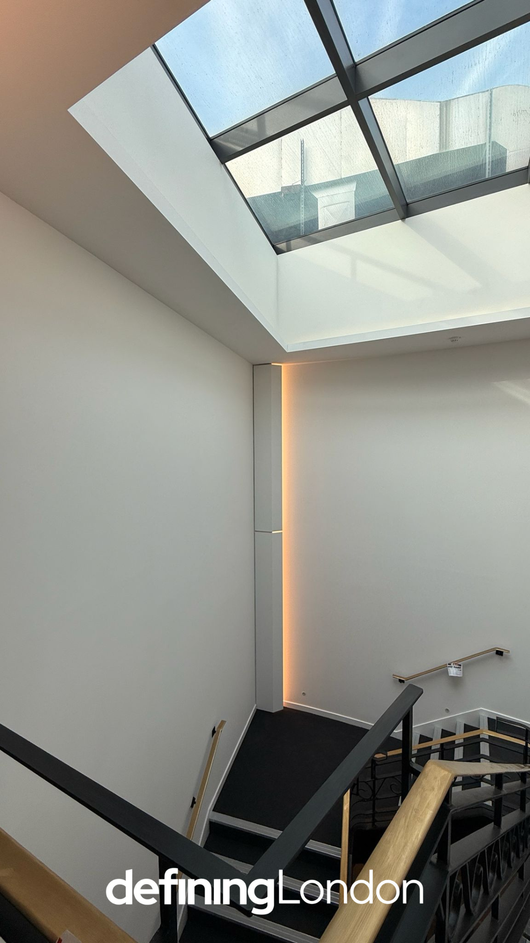 Interior view of a modern building staircase with a large skylight overhead, white walls, a black railing with gold accents, and a vertical strip of warm lighting.