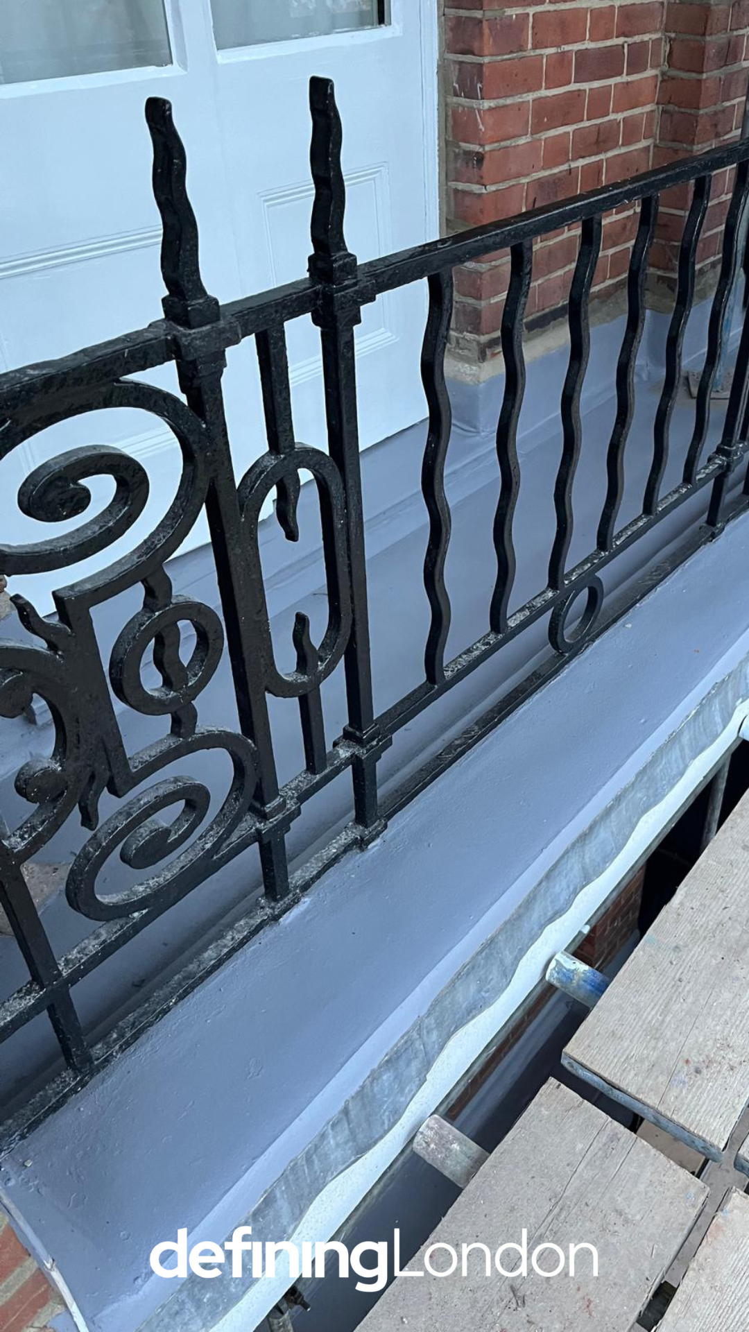 Close-up of a black wrought iron balcony railing with decorative swirls and vertical wavy bars in front of a brick wall and a blue door.