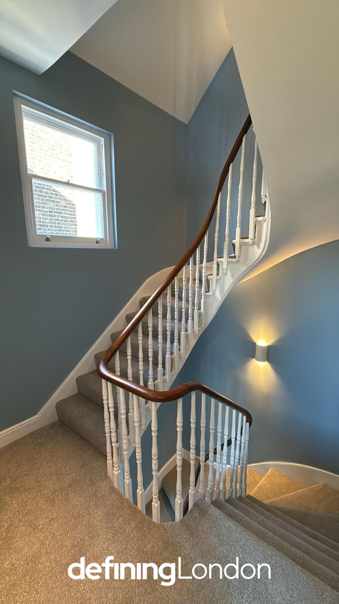 Interior staircase with beige carpet, white balustrades, and wooden handrail, against blue walls with a wall-mounted light fixture, next to a window with a view of brick exterior wall.