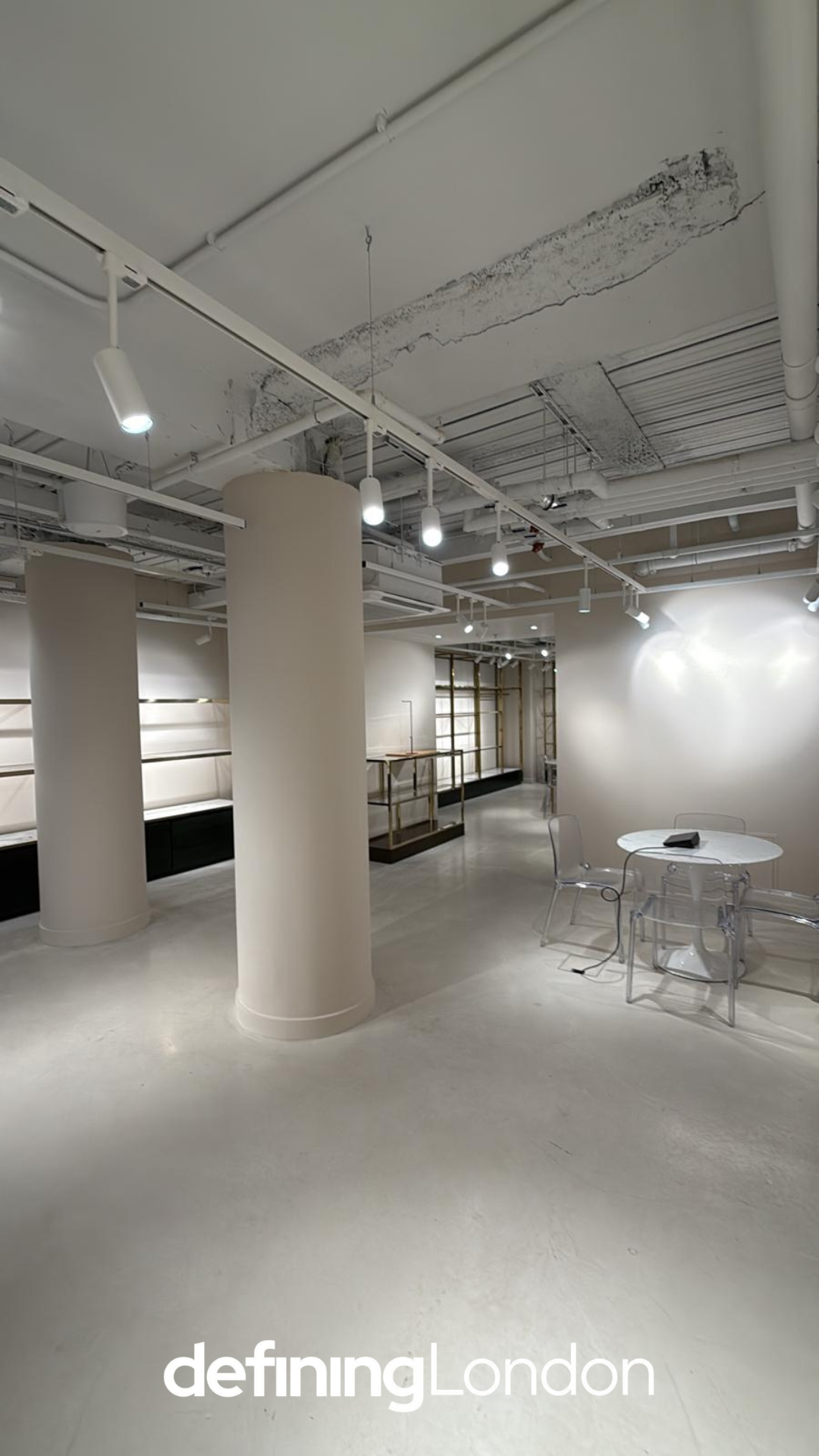 Empty modern retail space with white walls, large white pillars, and unfinished ceiling with exposed pipes and wiring, featuring a small round table with transparent chairs, and shelving units in the background. Text overlay says "definingLondon."