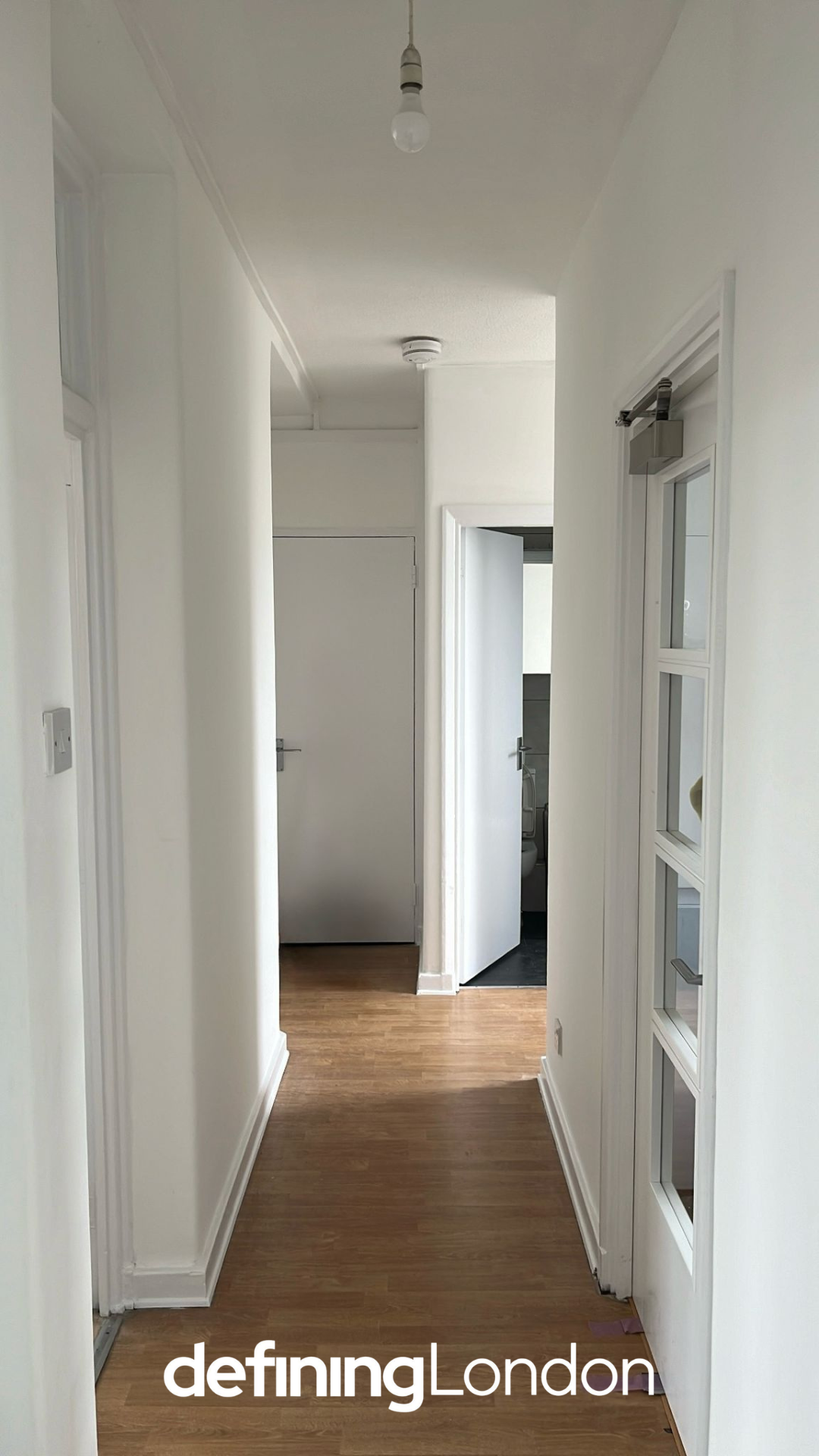 A hallway in a residential space with white walls, wooden floor, a light bulb hanging from the ceiling, and doors leading to a bathroom and another room, with a small glass display case on the right.