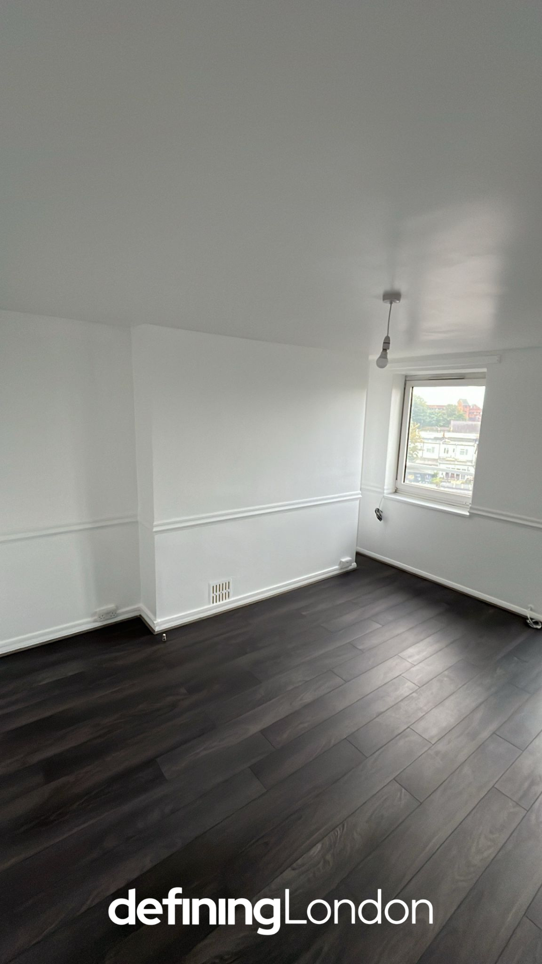 Empty room with white walls, dark hardwood floor, and a window showing a cityscape.