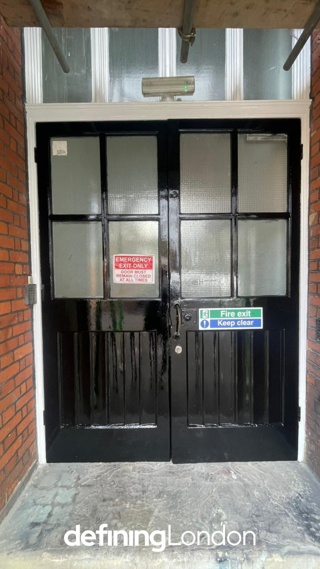 Black fire exit door with glass panels, emergency exit and fire safety signs, brick and glass exterior wall, concrete ground, and metal scaffold above.