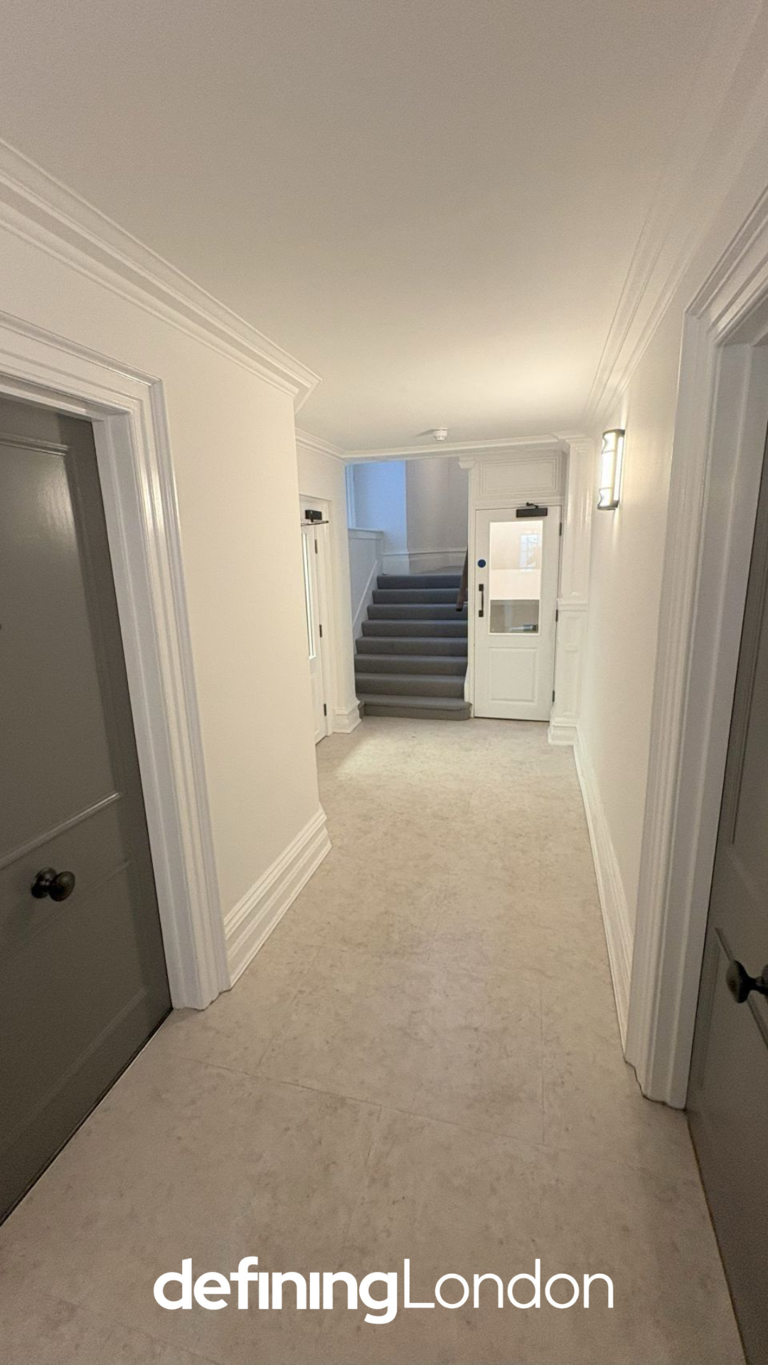 A hallway in a building with white walls and beige tile flooring, leading to a staircase with gray carpeted steps. There are several doors along the hallway, and a wall-mounted light fixture on the right side. The stairs go up to an unseen second floor.