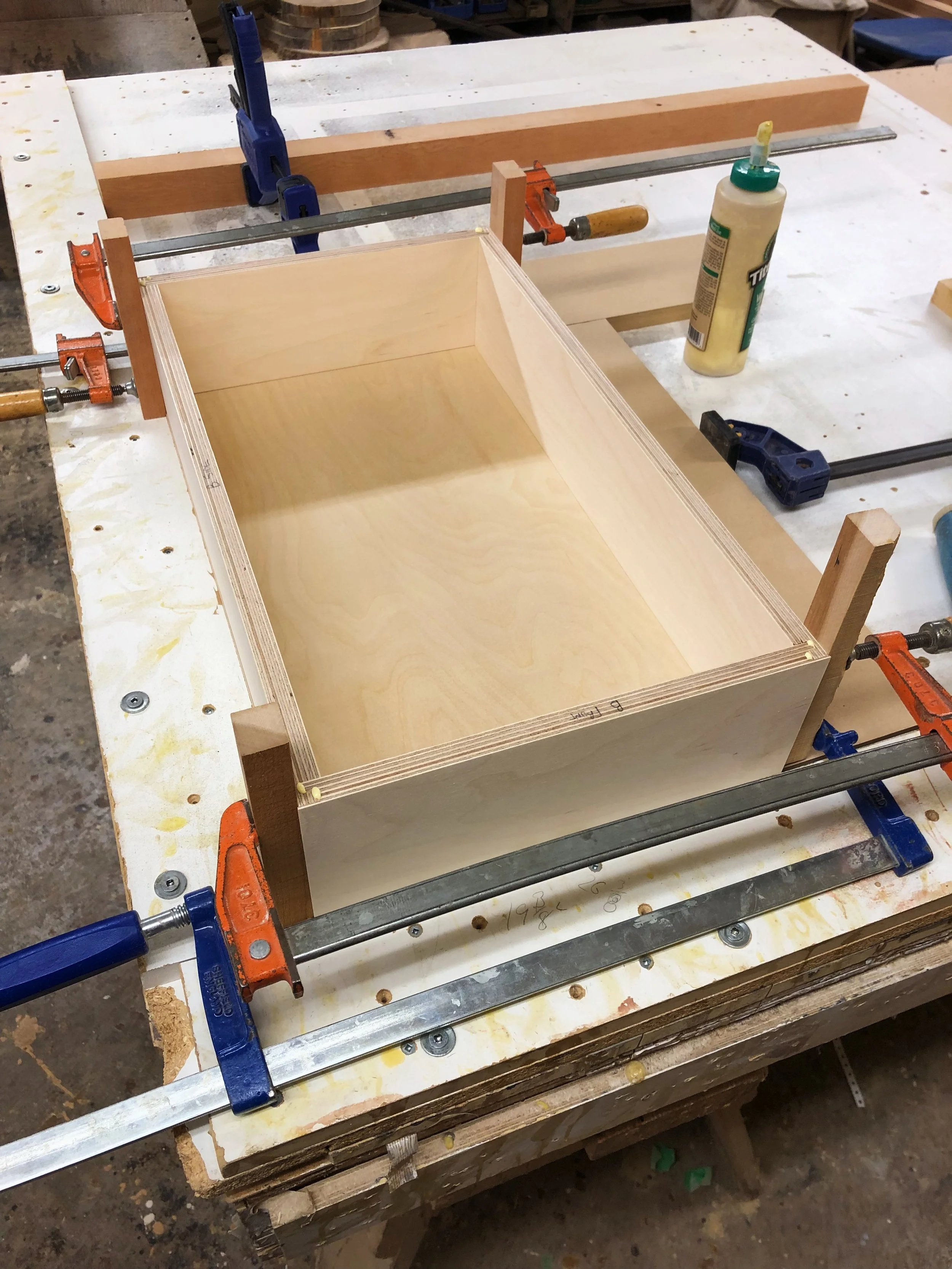 A woodworking project in progress with a partially assembled wooden box on a workbench, clamps securing the sides, and glue bottles nearby.