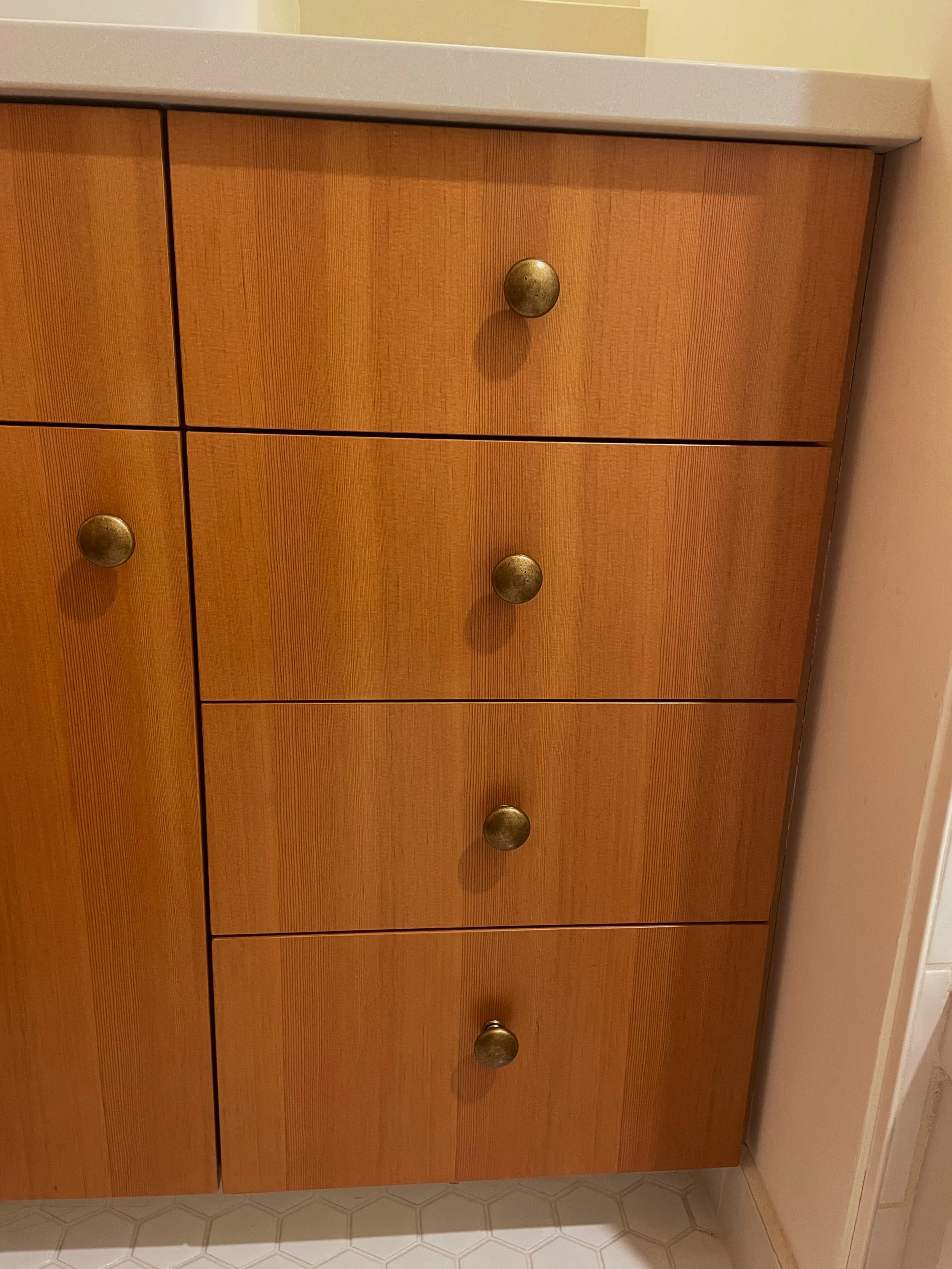 Wooden kitchen cabinet with six drawers, each with a round metal knob, situated against a tiled wall and topped with a light-colored countertop.