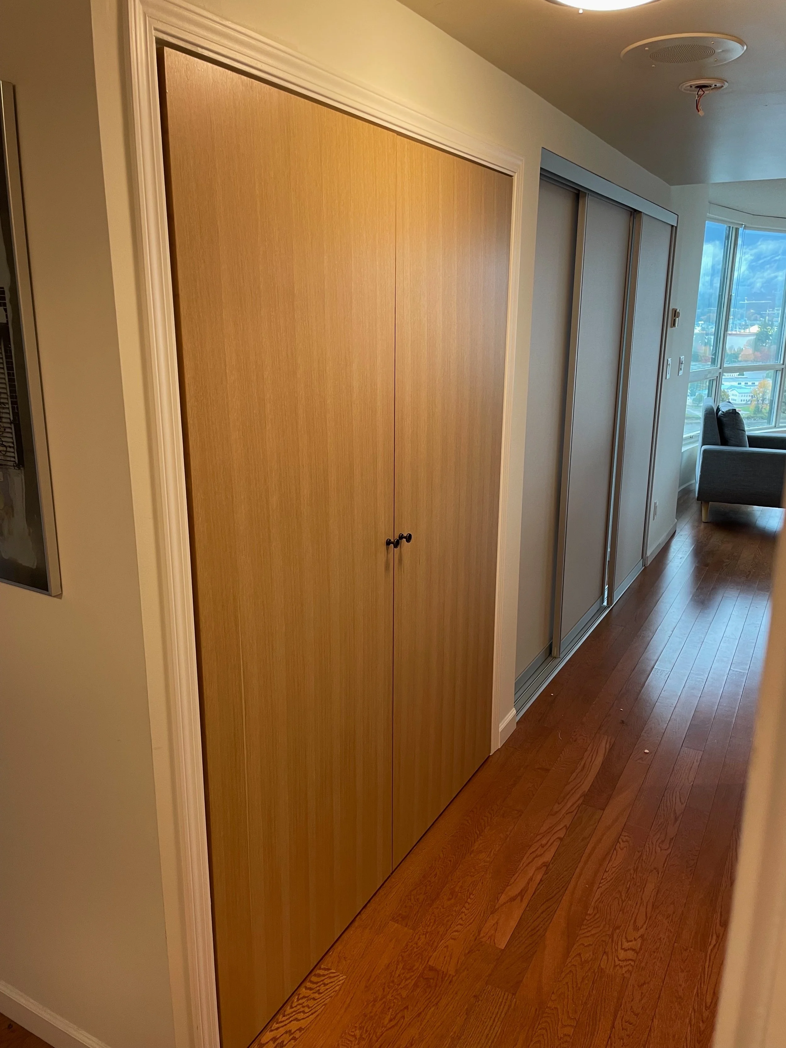 Wooden closet doors in a hallway with hardwood flooring and a large window at the end.