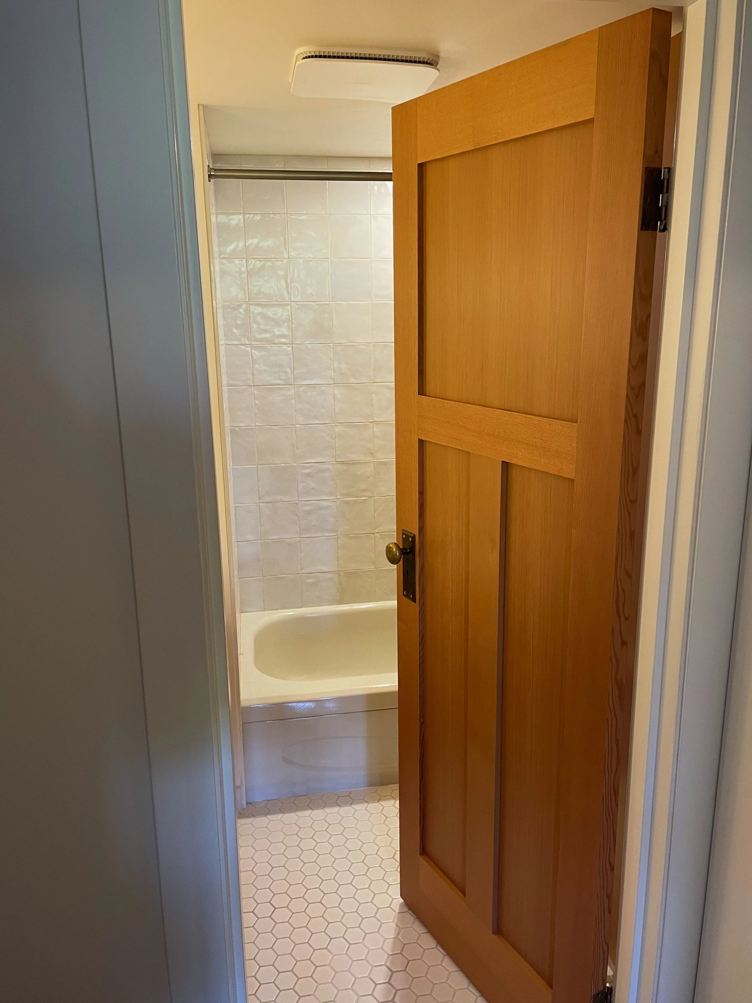 Open wooden bathroom door revealing a bathtub, tiled wall, and hexagonal floor tiles.