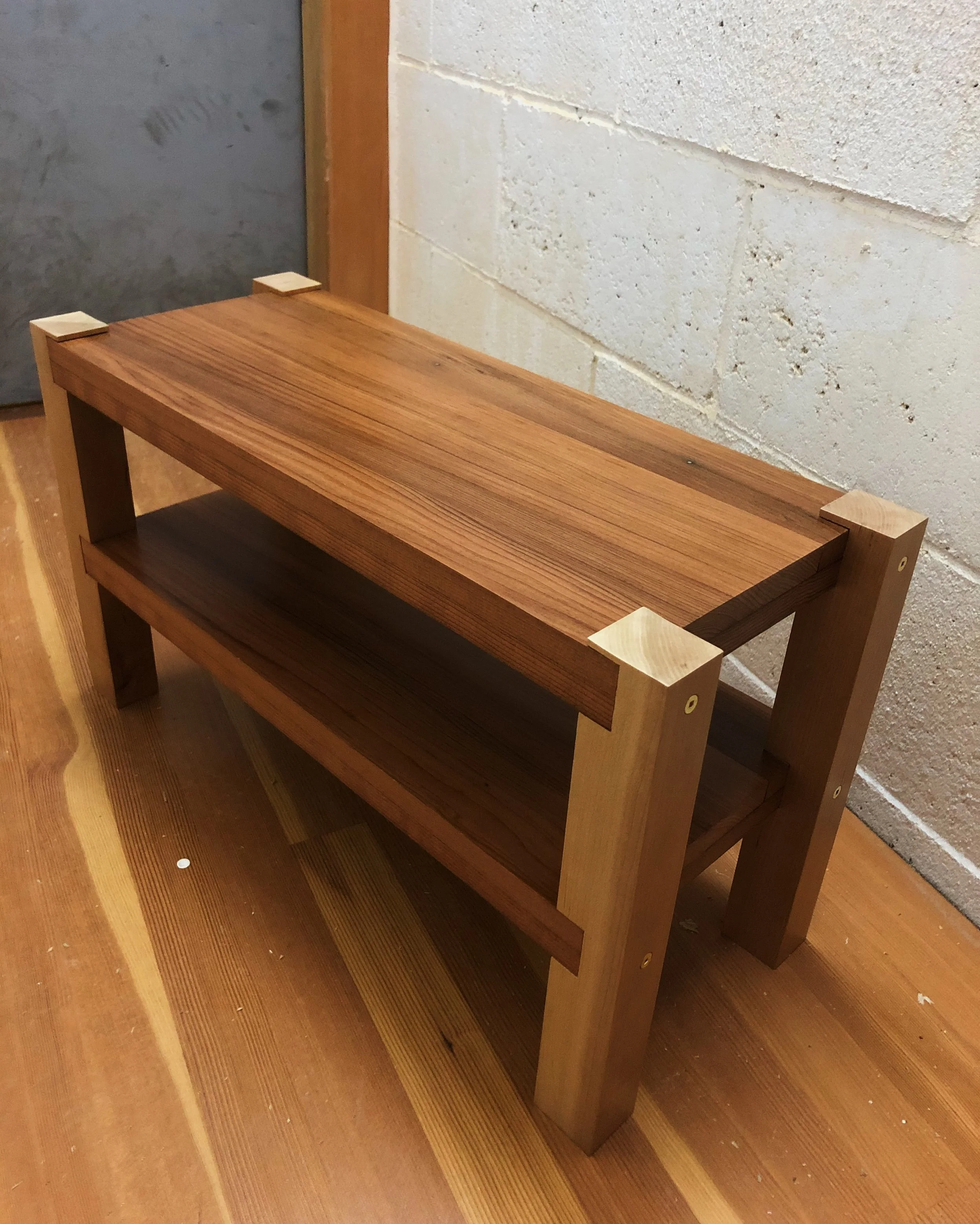A wooden two-tier bench with a natural finish, placed on a wooden floor near a white brick wall.