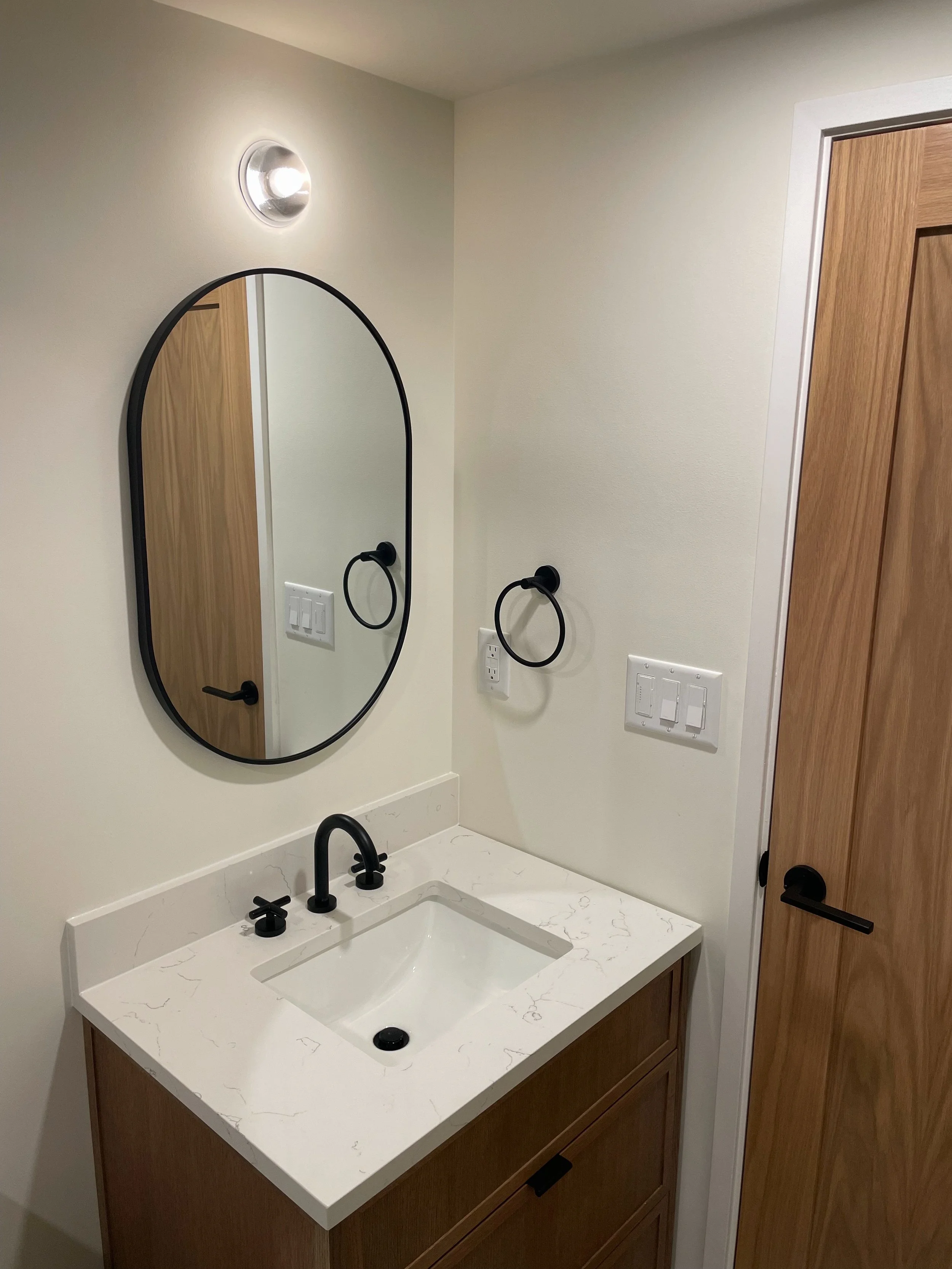 Bathroom vanity with white marble countertop, undermount sink, black faucet, oval mirror, black towel ring, light switch panel, and wooden door.