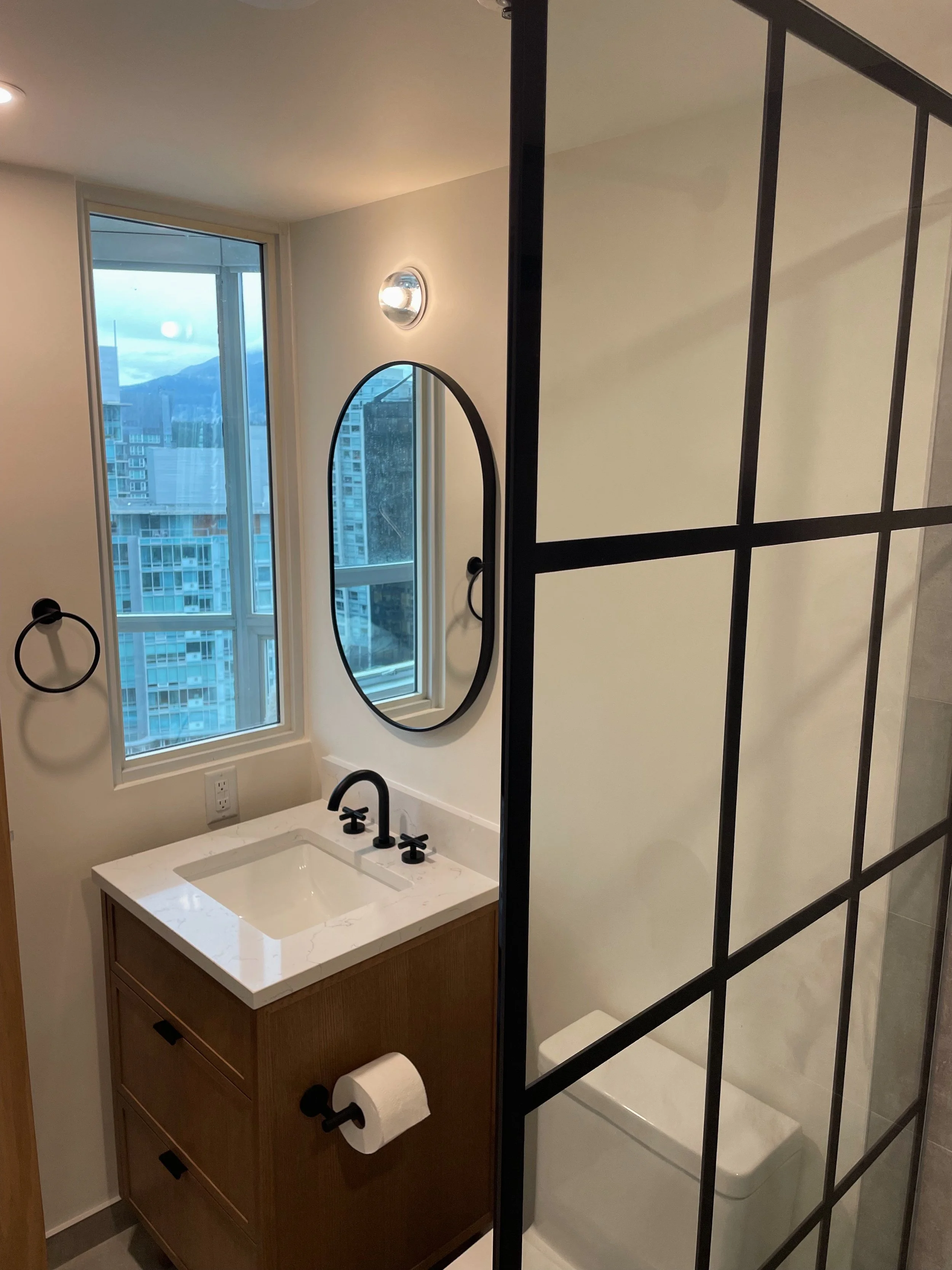 Modern bathroom vanity with a black faucet and oval mirror, next to a window with city view, separated by a black-framed glass shower wall.