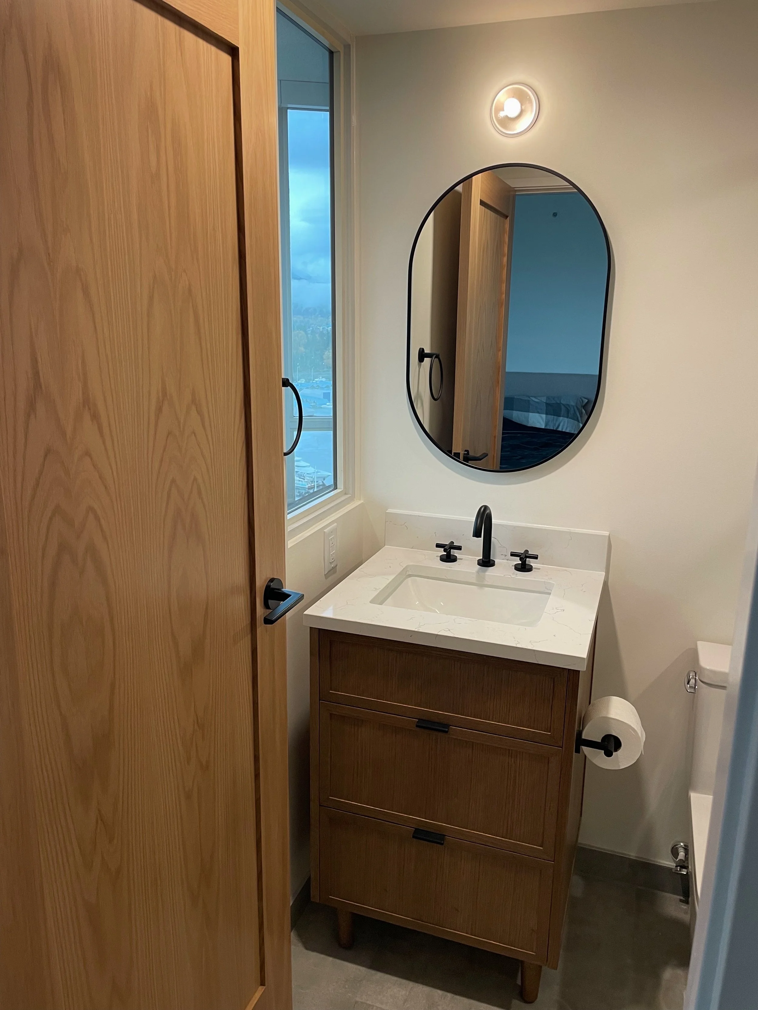 Small bathroom with a wooden door, a white vanity with a marble countertop, a black faucet, an oval mirror, a wall-mounted light, a window showing a cloudy sky, and a toilet paper roll on the side.