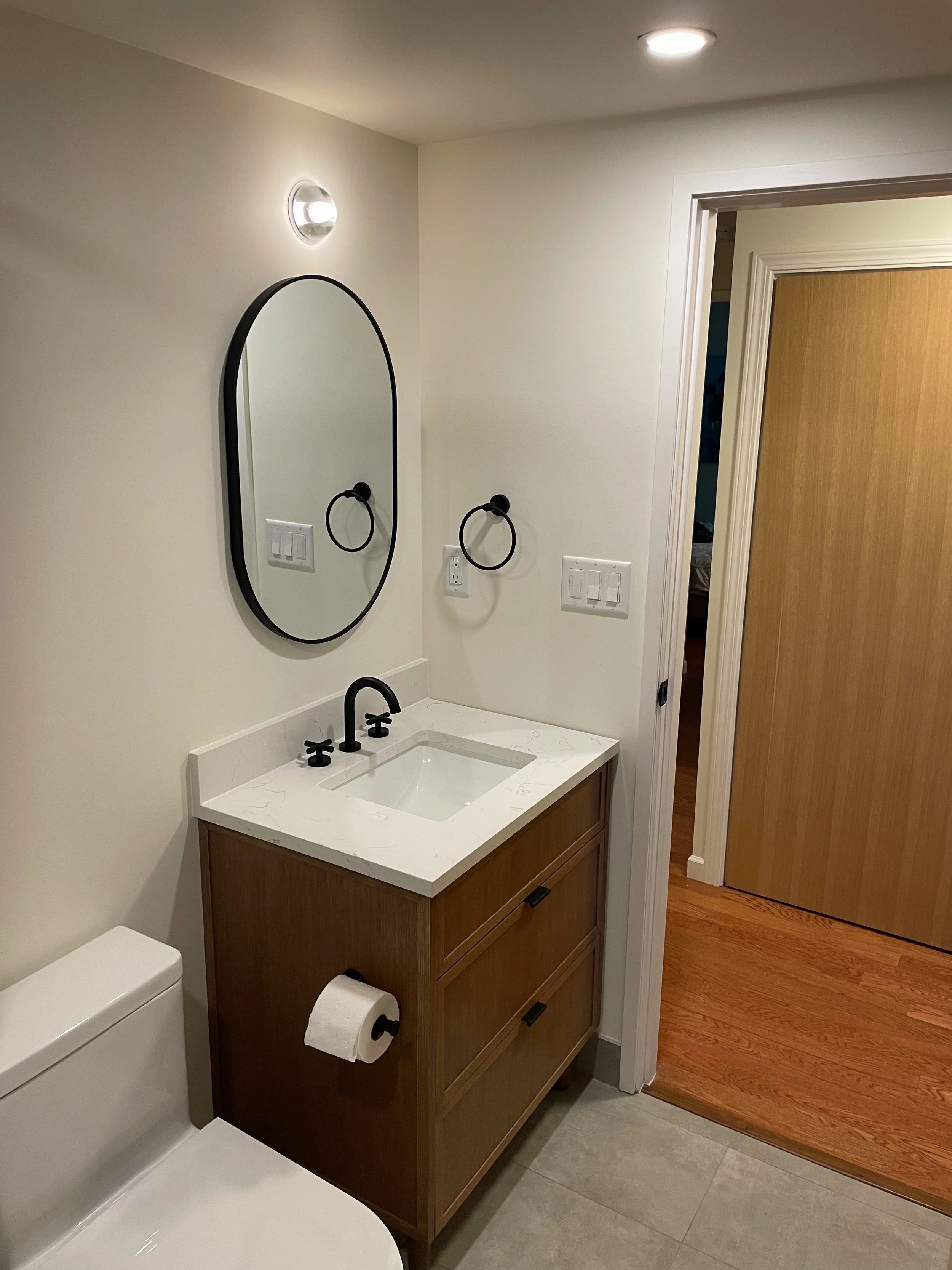 Bathroom with a wooden vanity, white sink, black faucet, oval mirror, wall-mounted towel ring, toilet paper holder, and a toilet, with a wooden floor and a sliding door.