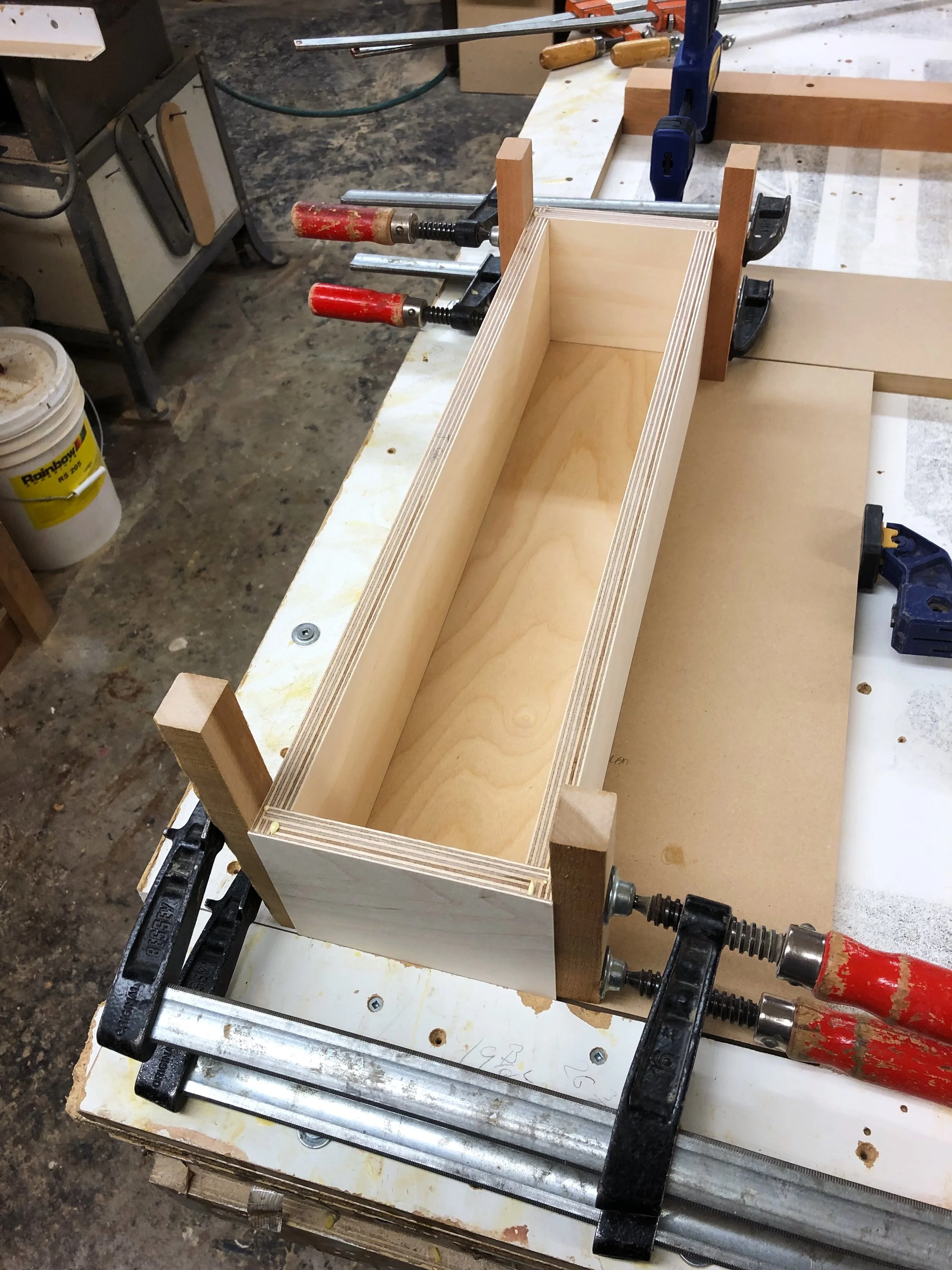 A wooden drawer being assembled and clamped on a workbench in a woodworking shop.