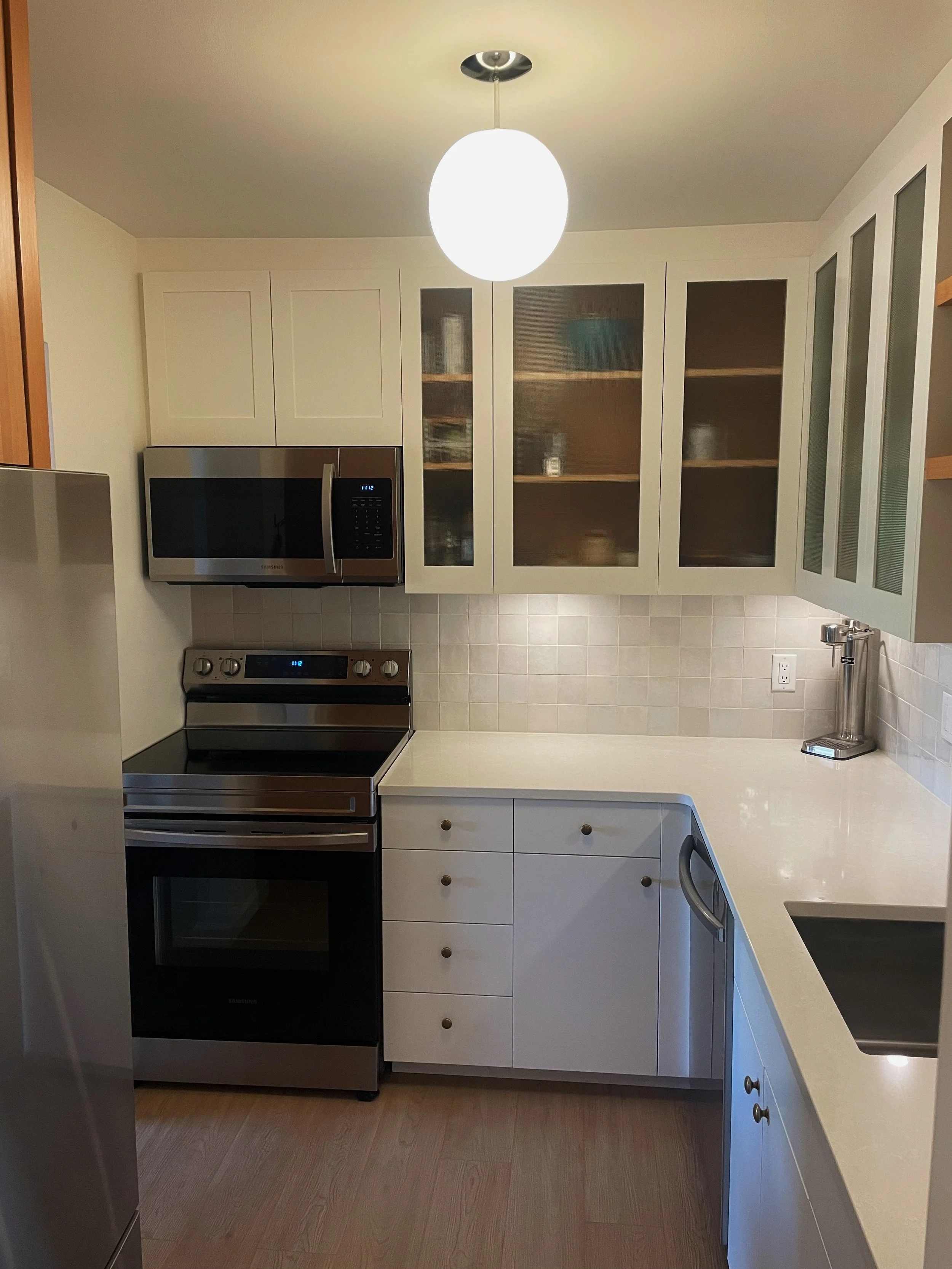 Modern kitchen with white cabinets, white countertops, stainless steel stove and microwave, beige tile backsplash, and a bright ceiling light.