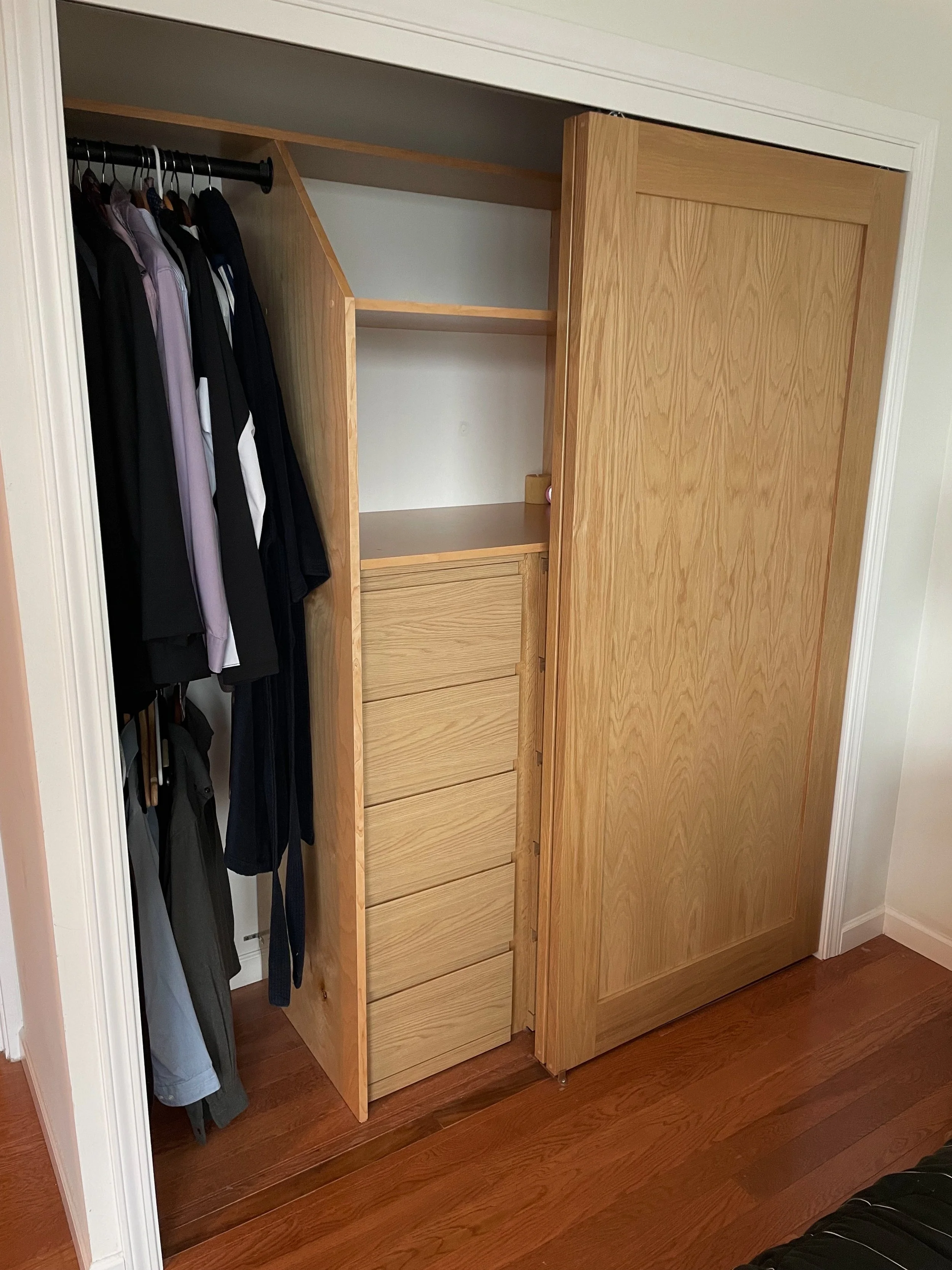 Open wooden closet with hanging clothes on the left and empty shelves and drawers inside.