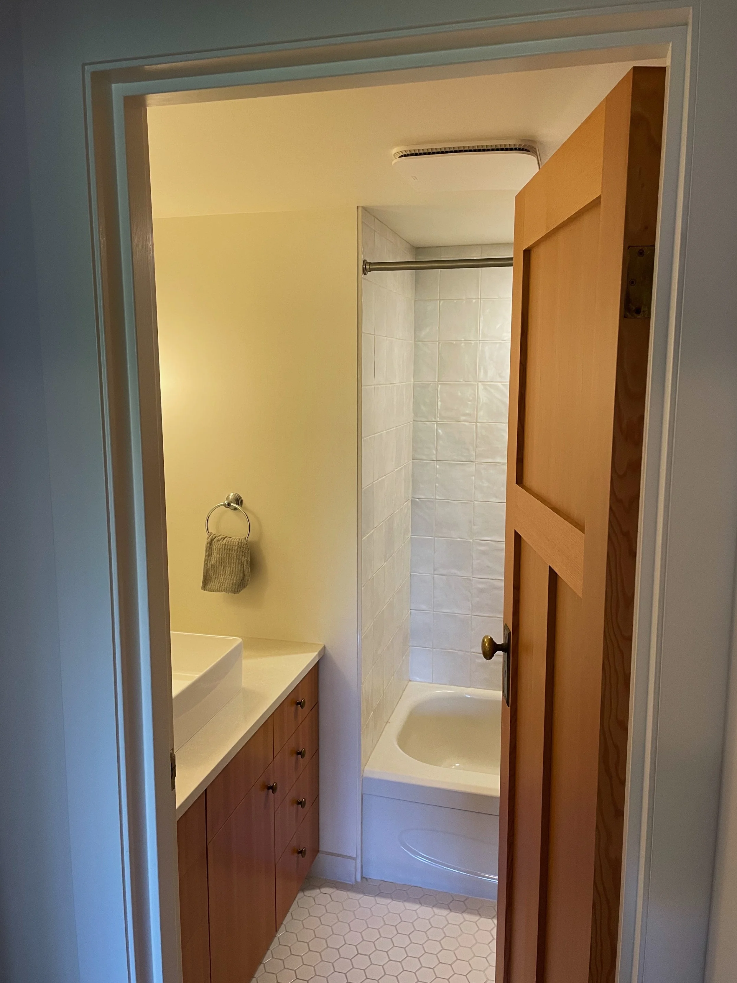A small bathroom with a wooden door partially open. Inside, there is a white sink with a wooden cabinet underneath, a towel ring with a towel, and a bathtub with white tiles on the wall.