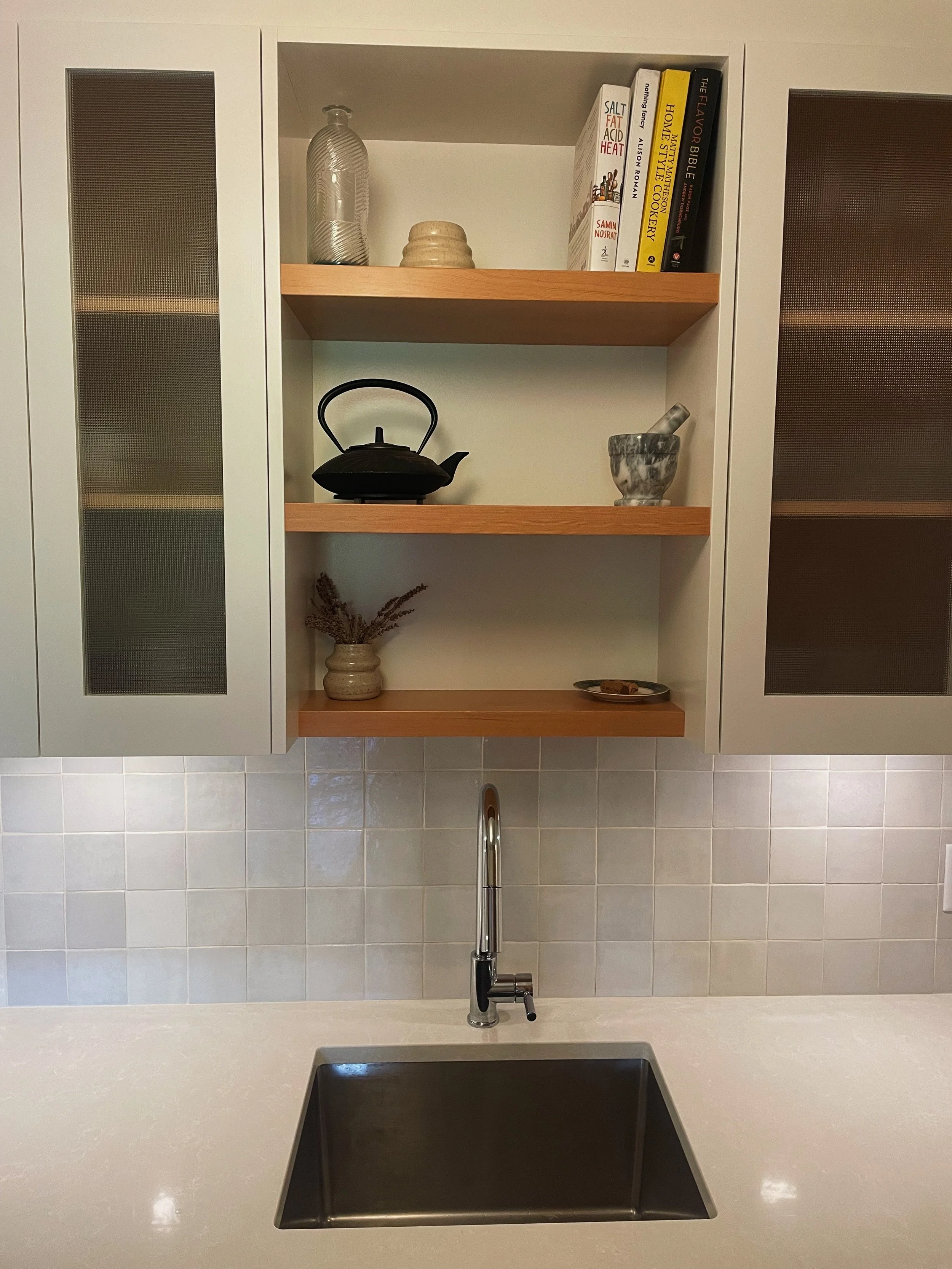 Kitchen with open wooden shelves holding glass jar, kettles, books, bowl, and small dish, with a stainless steel sink and faucet below.