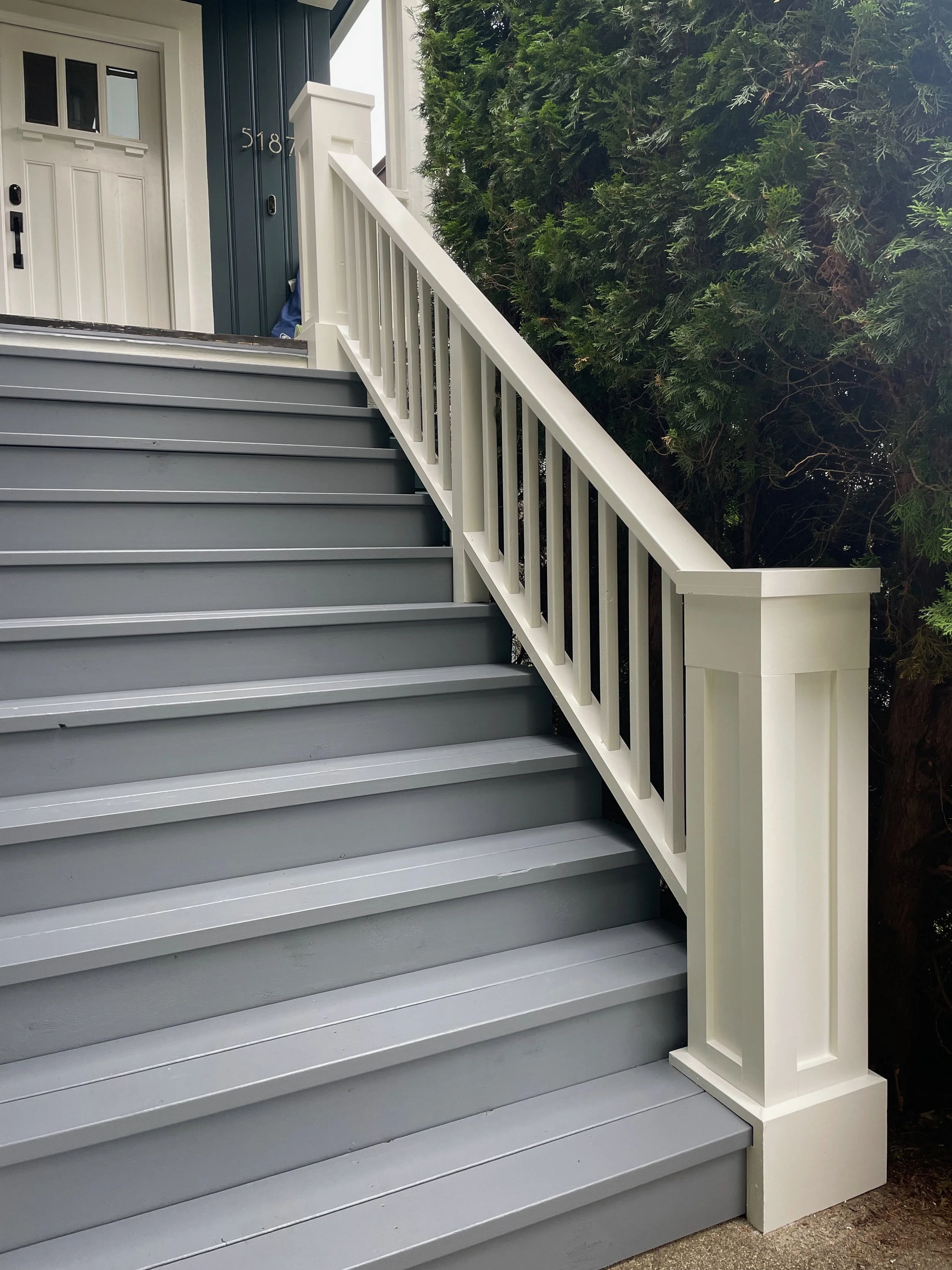 Close-up of exterior stairs with gray steps and a white railing leading to a front door, with a large green bush on the right side.