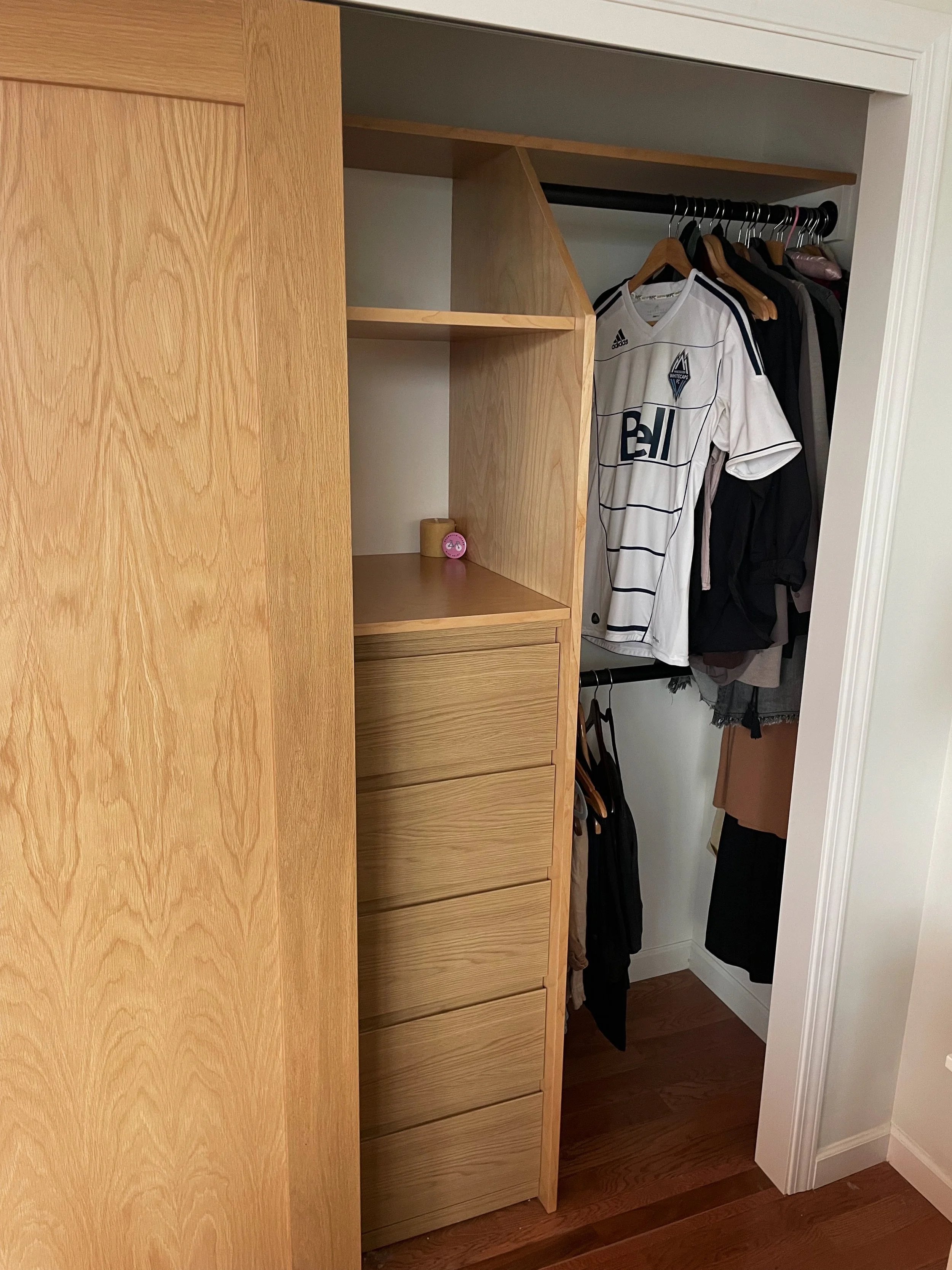 Open closet with hanging clothes, including a white sports jersey, and a built-in wooden dresser with six drawers. A small pink alarm clock is on a shelf inside the closet.