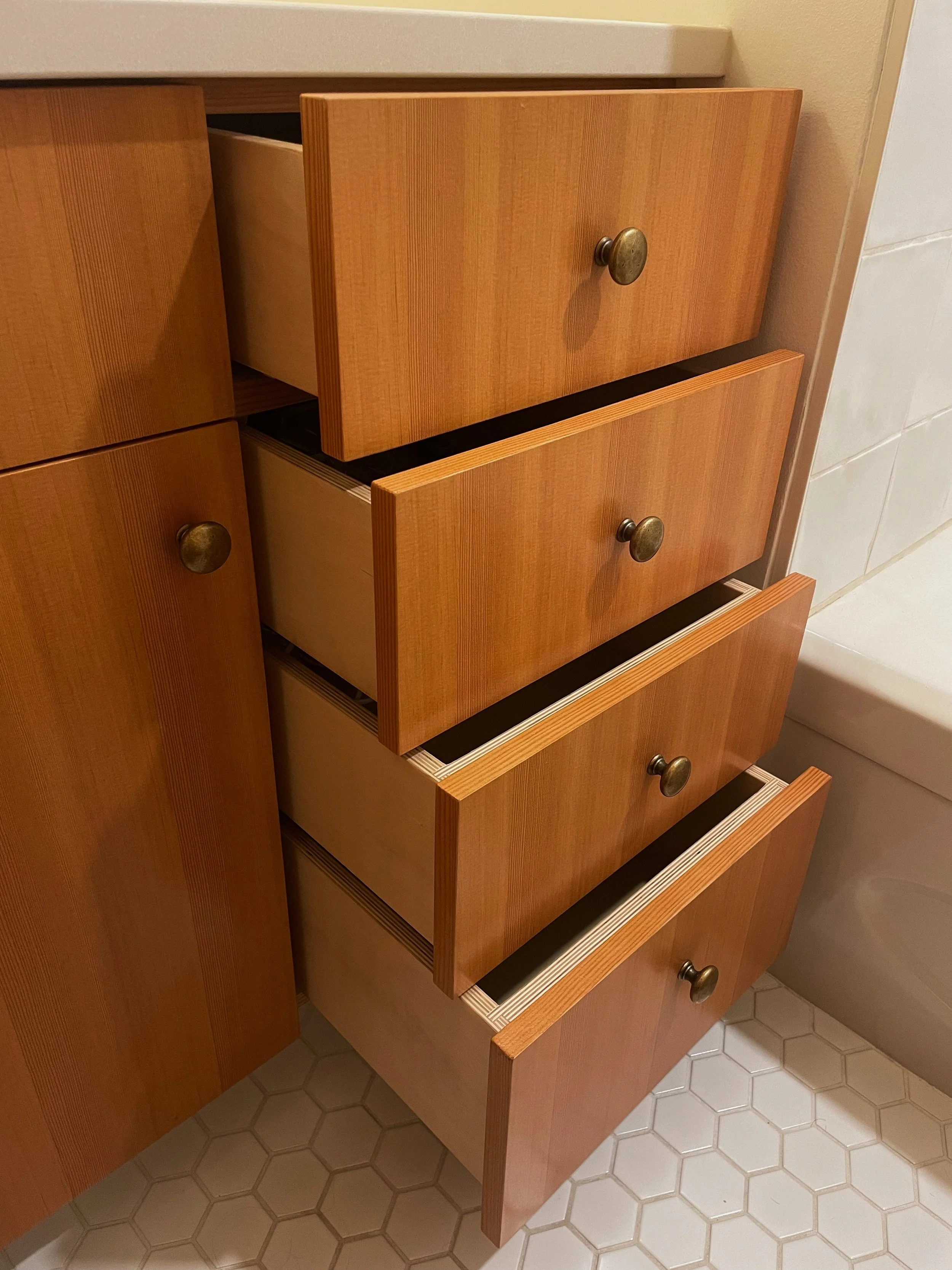 Four open kitchen drawers with wood finish, metal knobs, and white interior, partially pulled out next to a white tiled floor and beige wall.