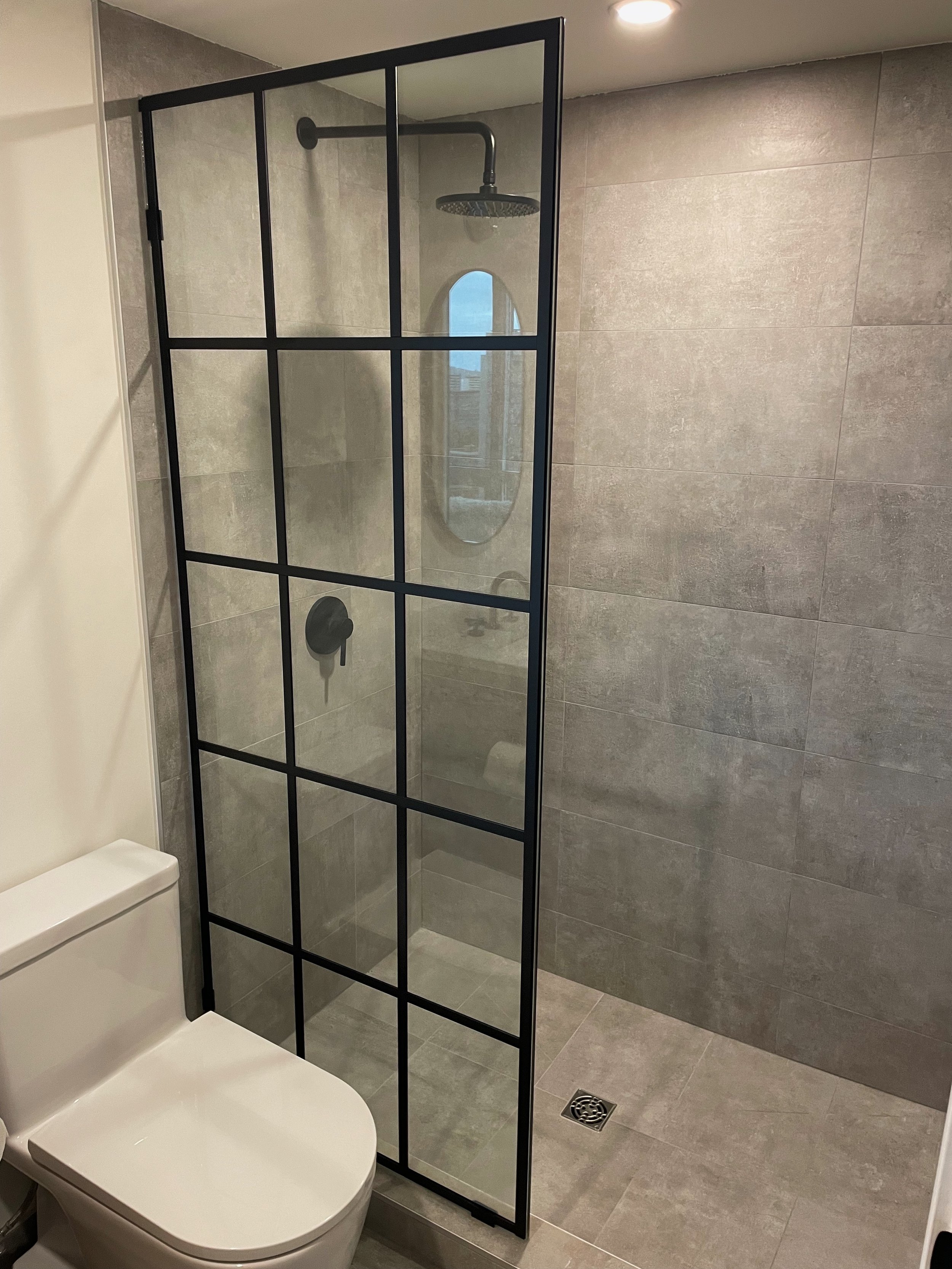 Modern bathroom with a walk-in shower enclosed by a black-framed glass partition, grey tiled walls and floor, a rain showerhead, and a small window.