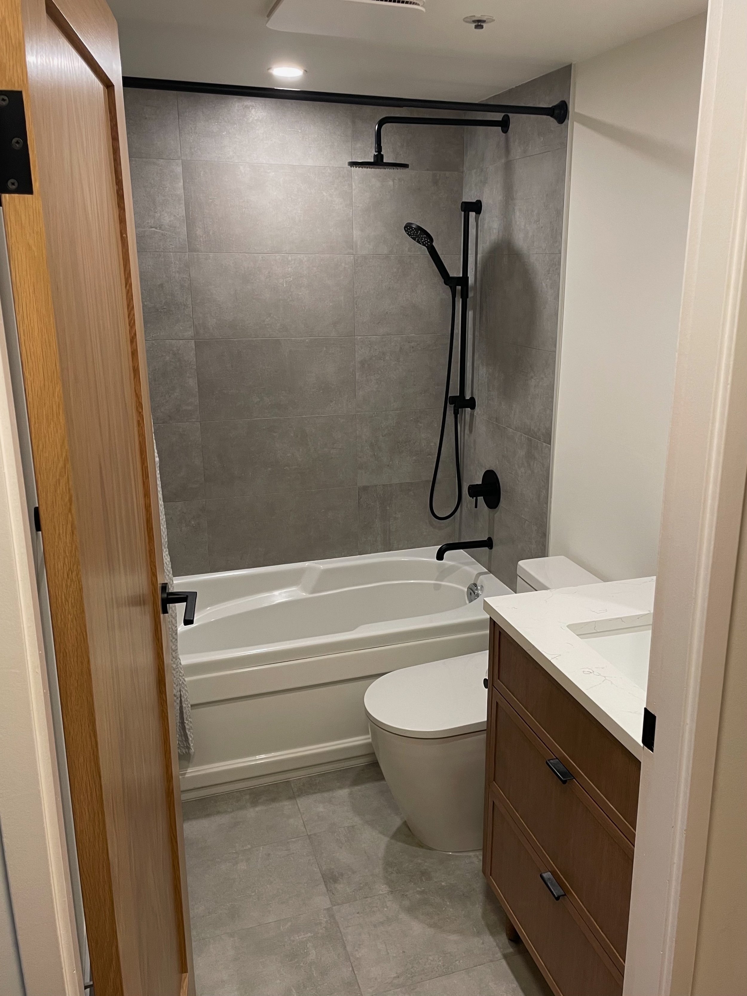 A small bathroom with a bathtub, black shower fixtures, a toilet, and a wooden cabinet with a white countertop sink.