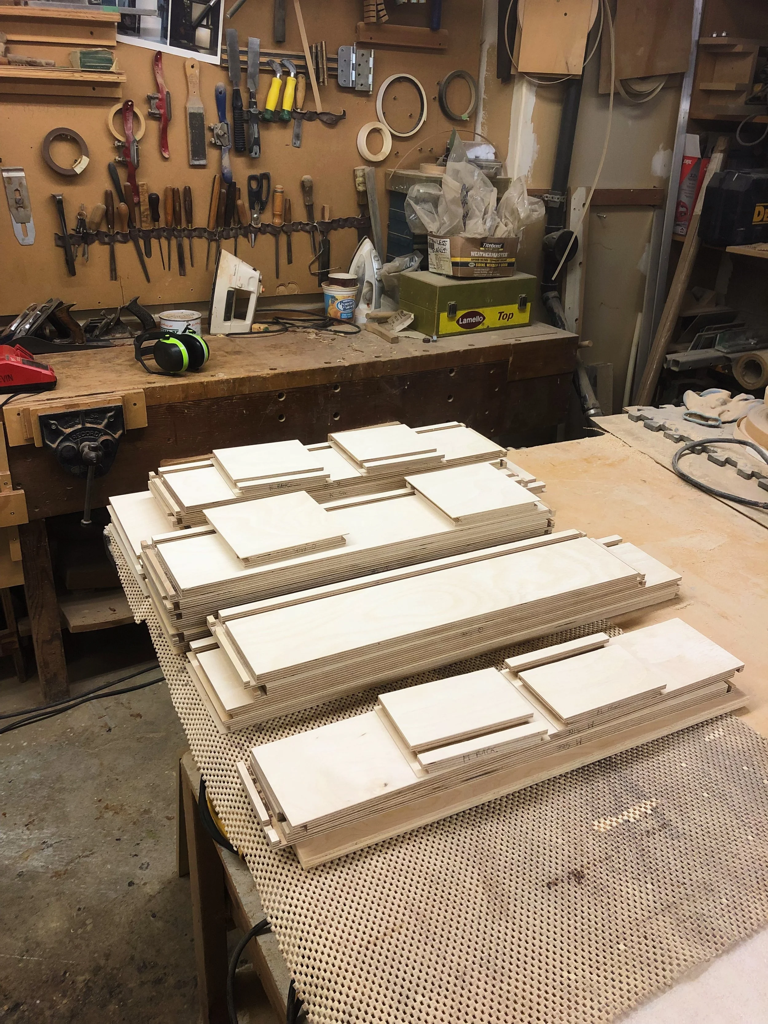 Stacks of unpainted wood pieces arranged on a woodworking workbench in a workshop, with tools and supplies hanging on the wall behind.
