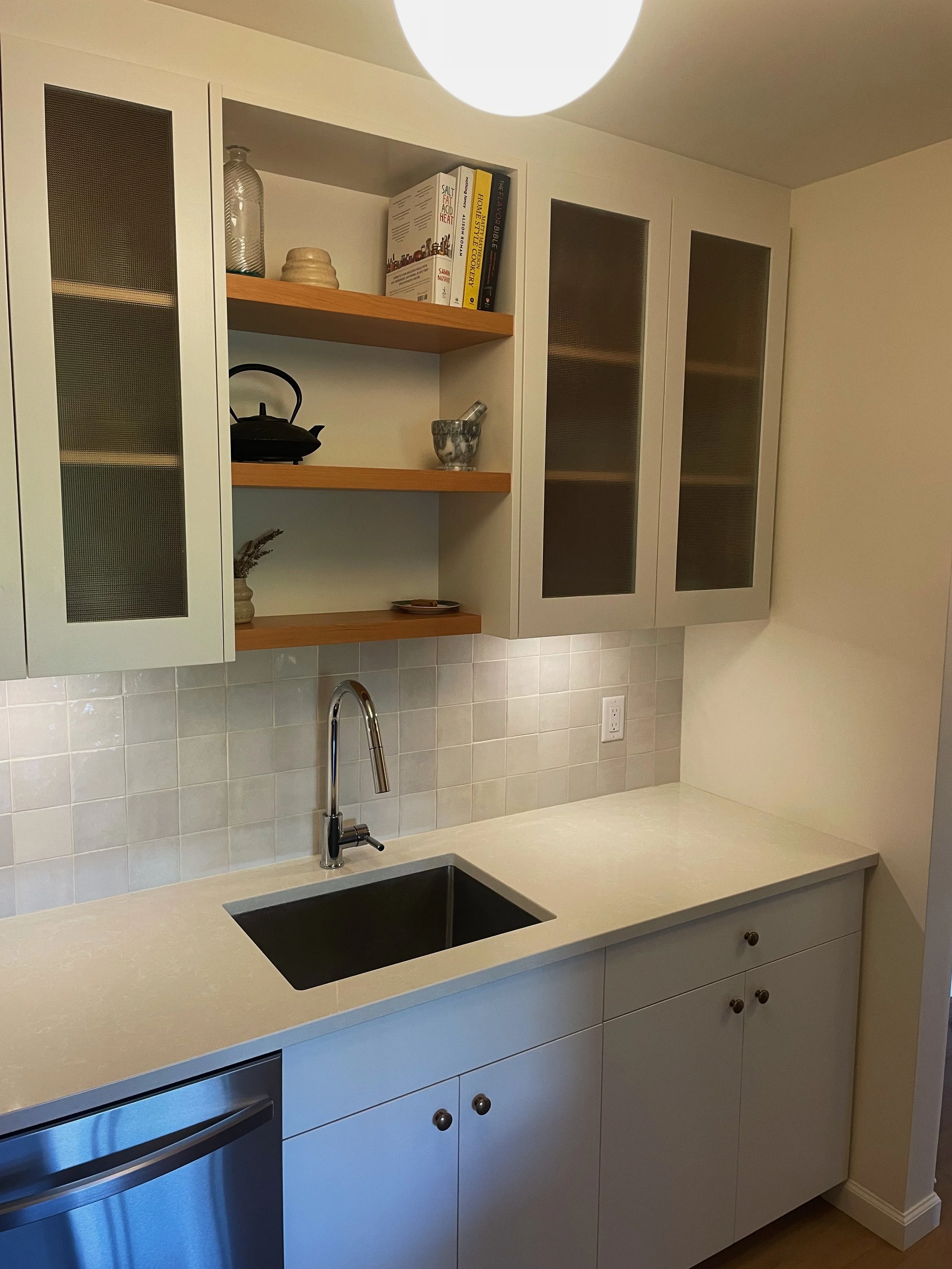Kitchen with white cabinets, a beige countertop, a black sink, upper cabinets with glass doors, open shelves with decorative items and books, and a reindeer ornament. There is a ceiling light and a tile backsplash.