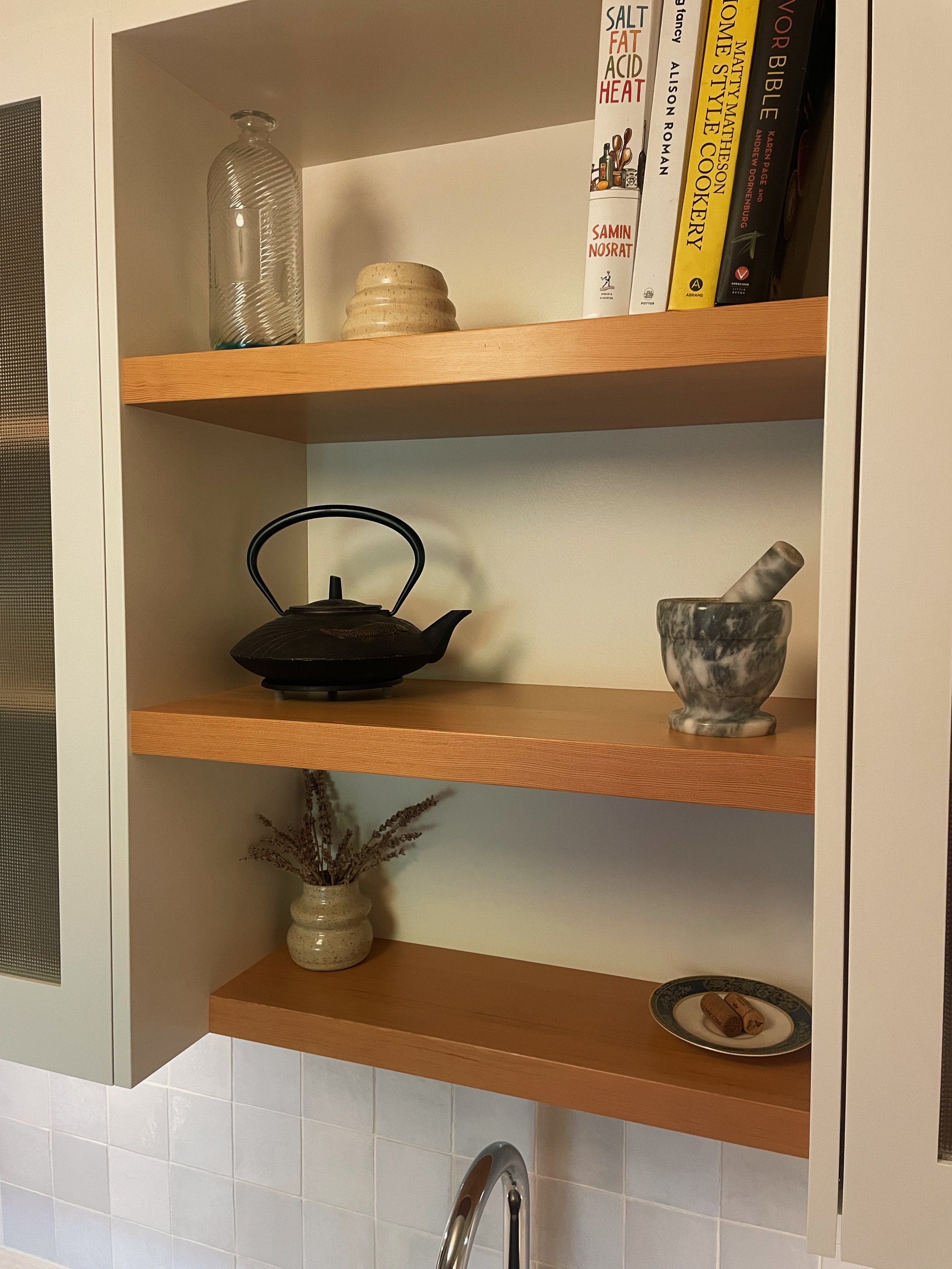 Open kitchen cabinet with three wooden shelves holding a glass bottle, ceramic dishes, books, a teapot, a marble mortar with pestle, a vase with dried plants, and a small plate with cinnamon sticks.