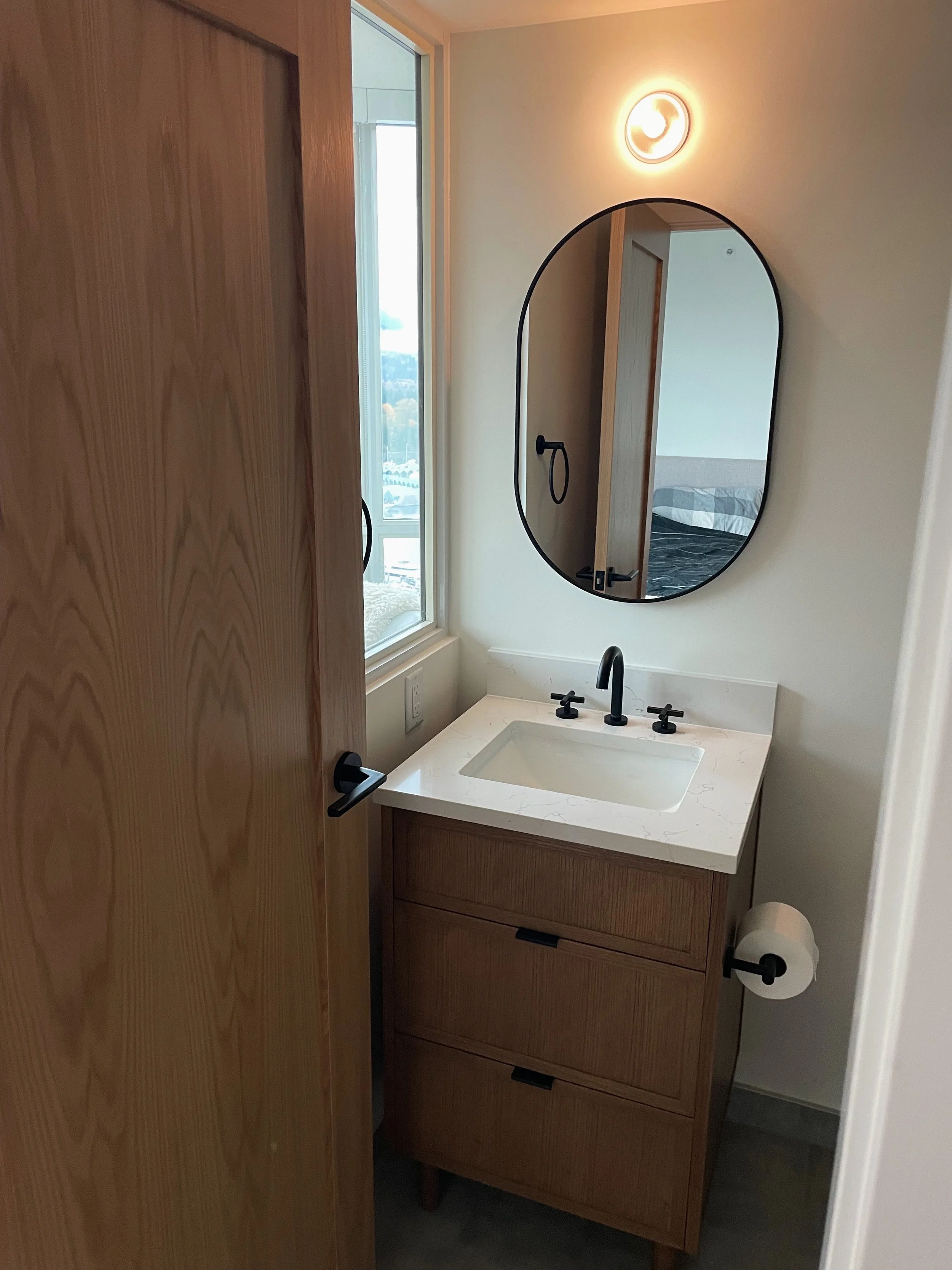 Small bathroom with a wooden vanity, white countertop, black faucet, oval mirror, wall-mounted light, and a window with a view outside.