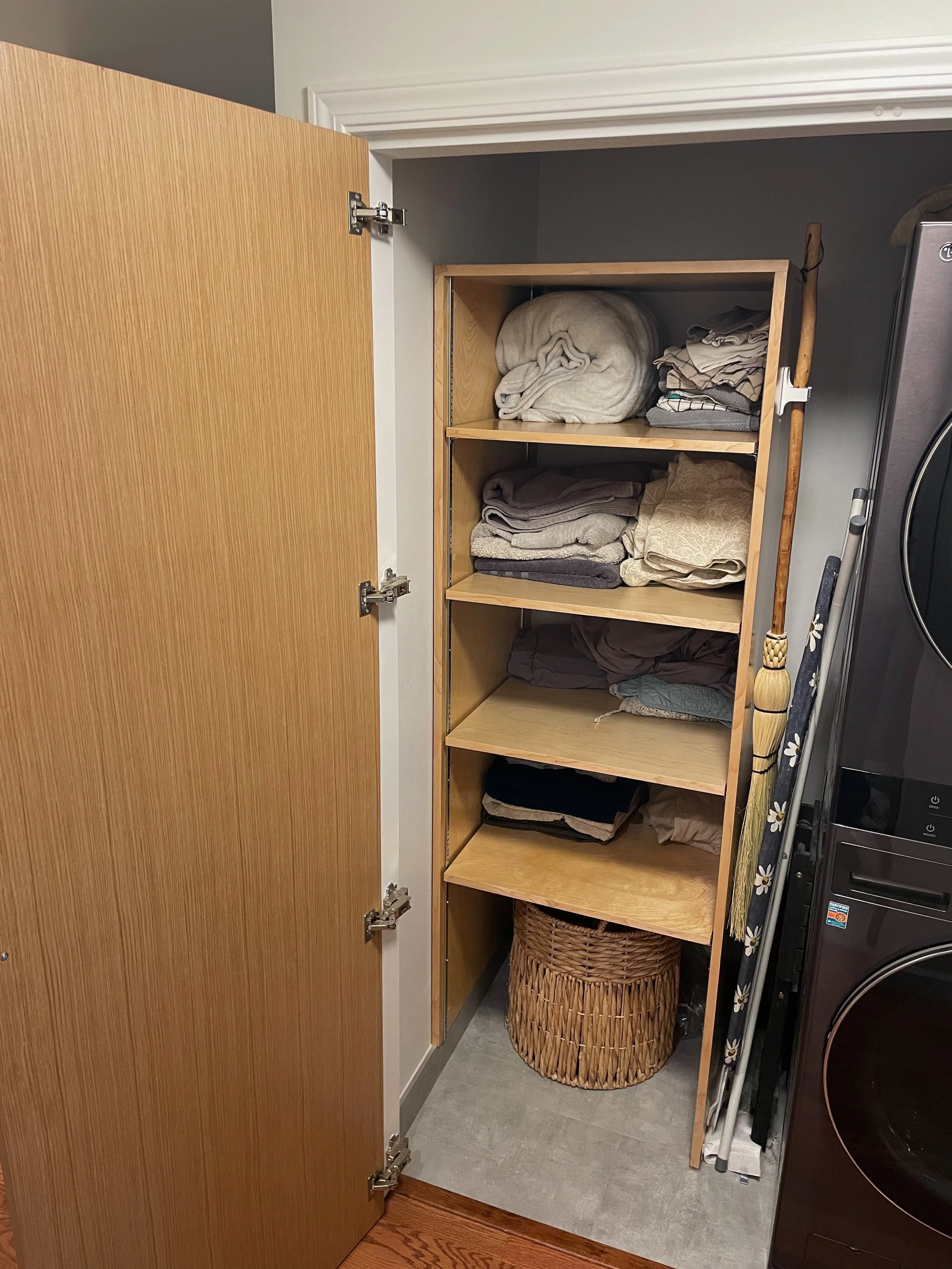 Open closet door revealing a wooden shelving unit with folded towels and linens on the shelves, a woven laundry basket underneath, a broom and vacuum cleaner beside the shelf, and laundry appliances to the right.