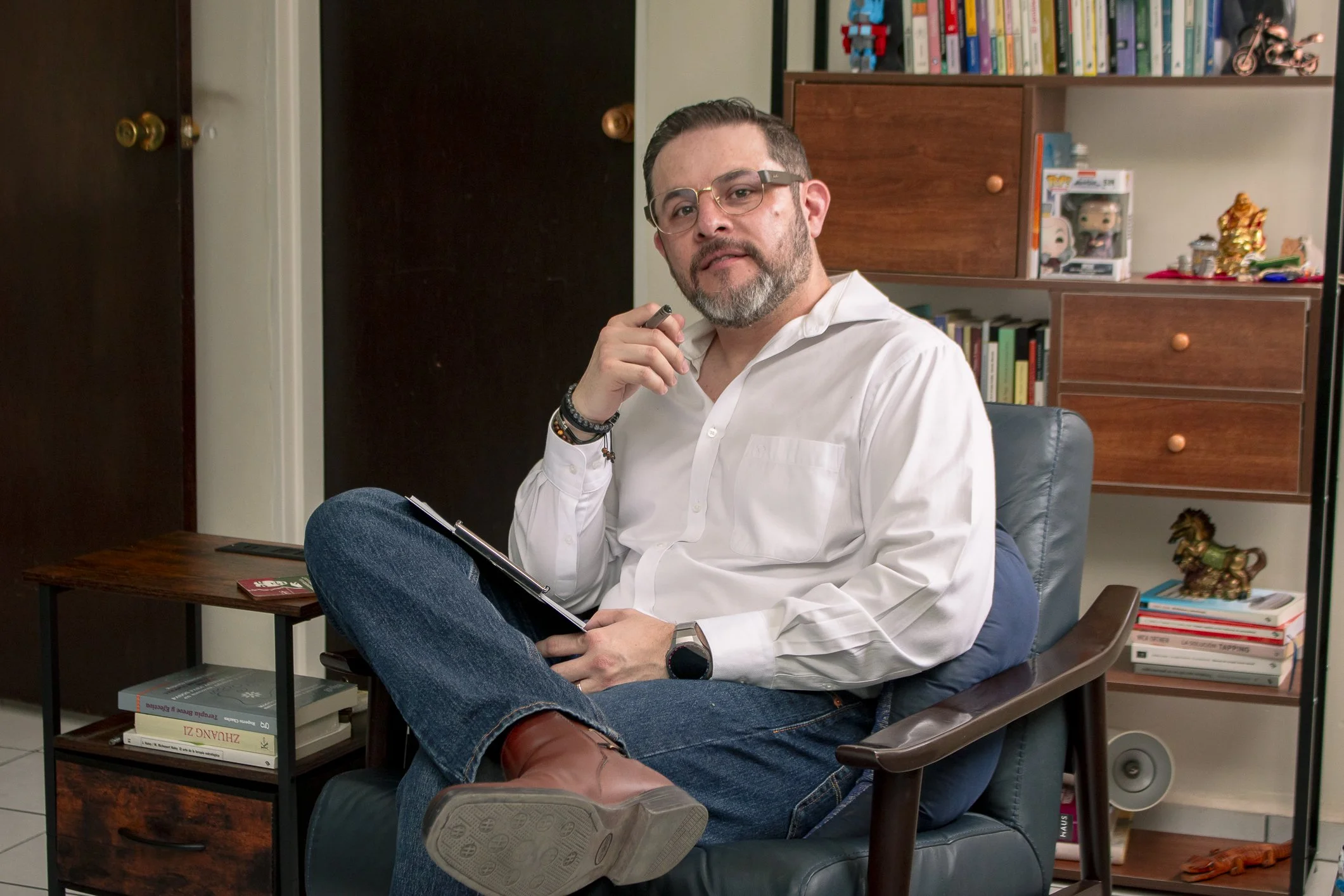 Psicologo Hombre con barba y gafas sentado en una silla de oficina, sosteniendo una pluma y un bloc de notas en una habitación con estanterías llenas de libros y decoraciones.