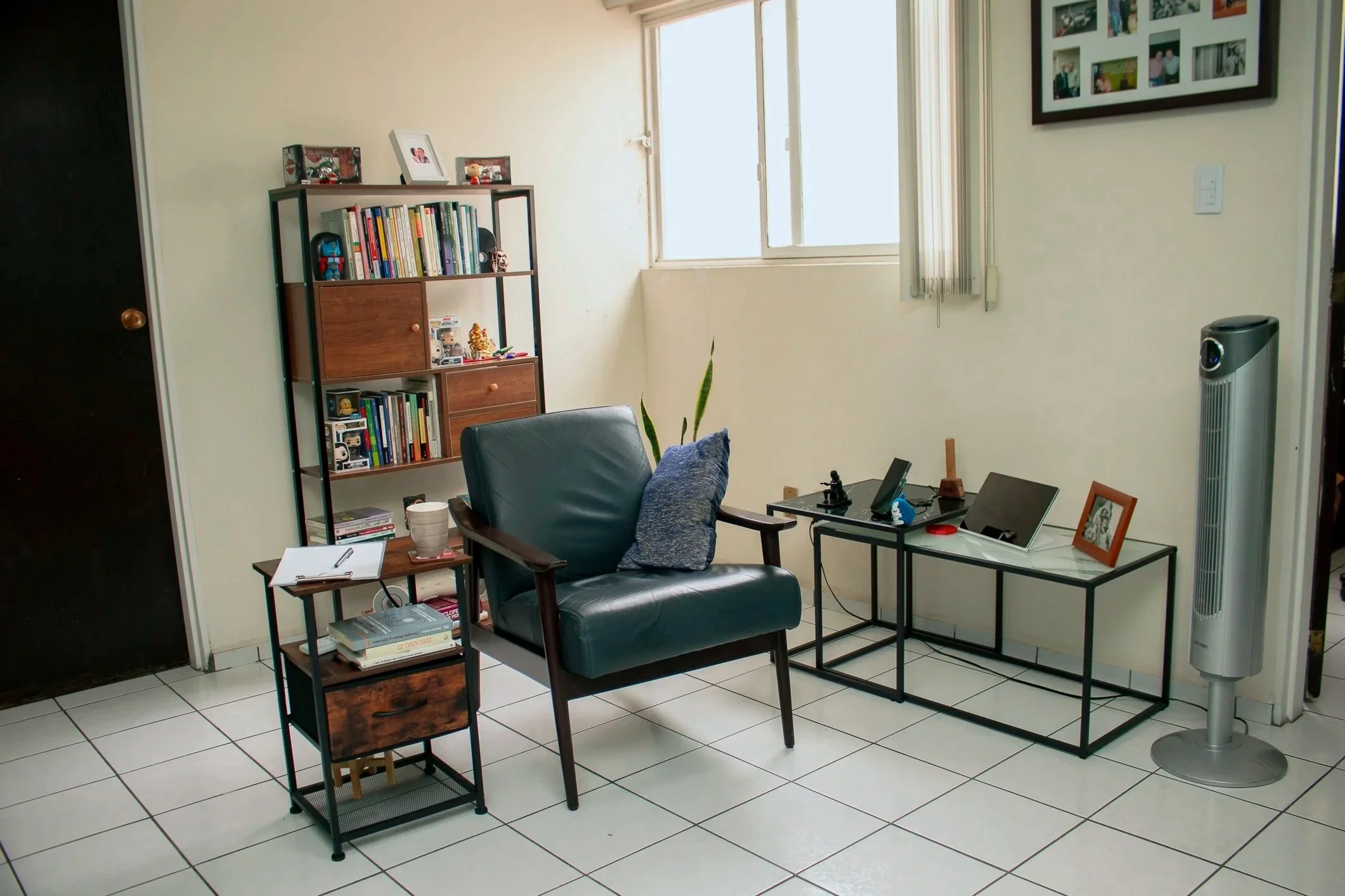 Consultorio, sala de estar con sillón de cuero, estantería de libros, mesa con laptop y objetos, ventilador de piso, y cuadros en las paredes.