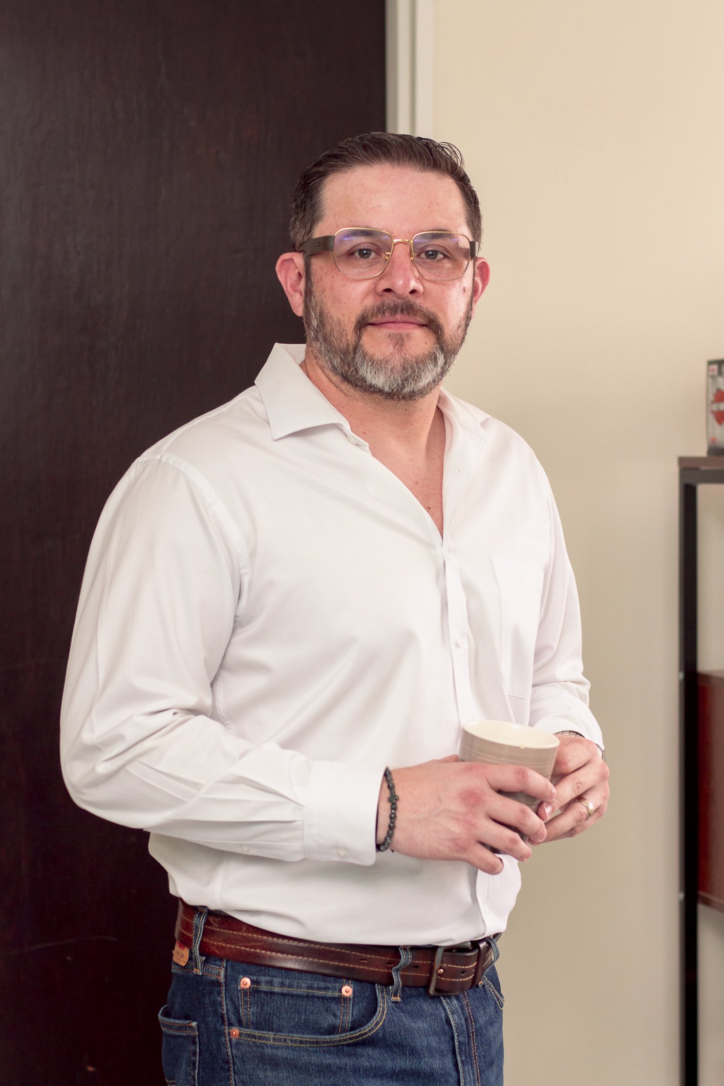 EL psicolgogo es Hombre con barba y gafas de montura gruesa, vestido con camisa blanca y jeans, sosteniendo un vaso de yogurt en un entorno interior.