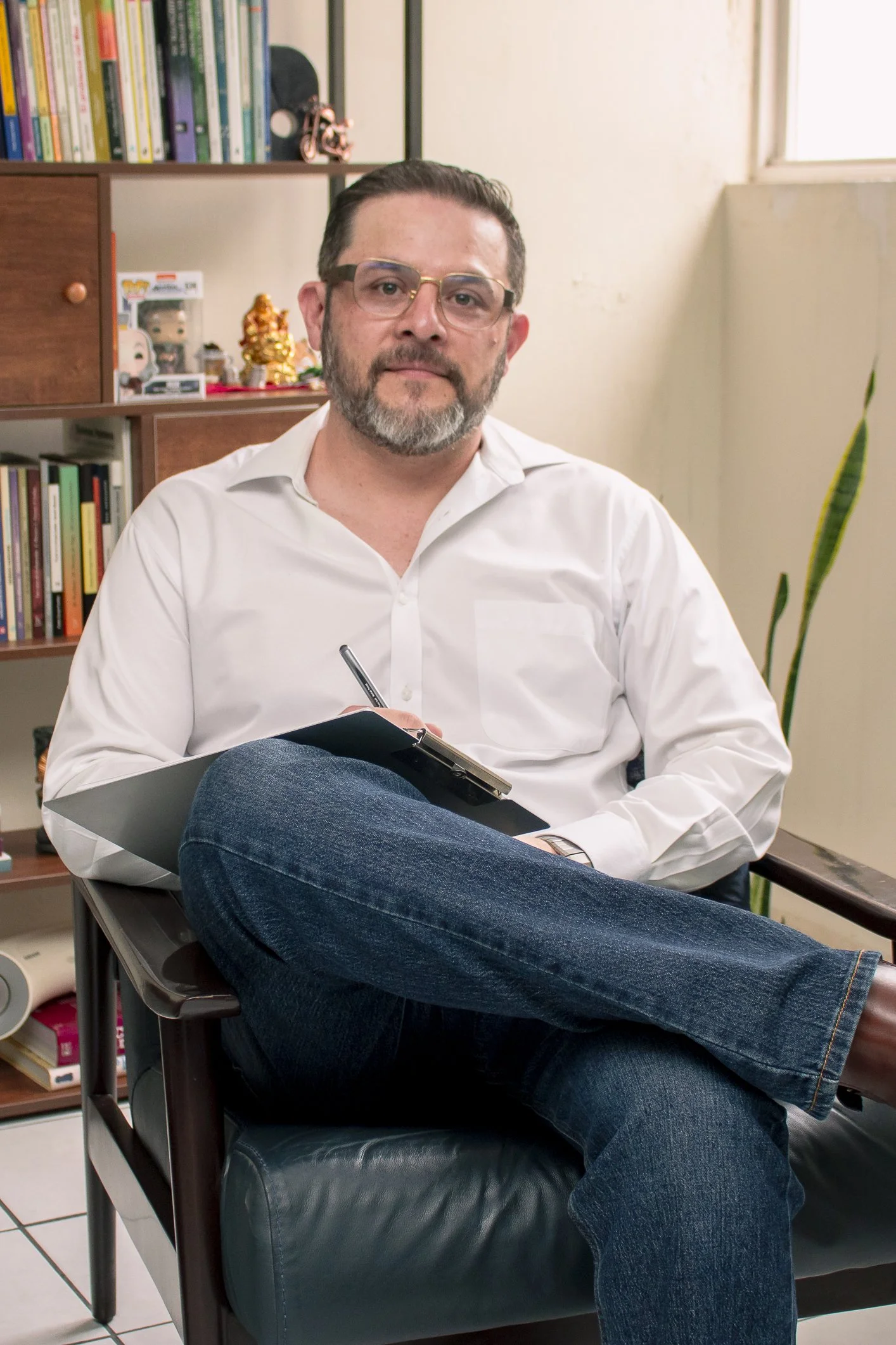 Psicologo, hombre con barba, gafas y camisa blanca siesta en una silla de oficina con un cuaderno y bolígrafo en las manos, fondo con estantería con libros y figuras decorativas, planta cerca de la ventana.