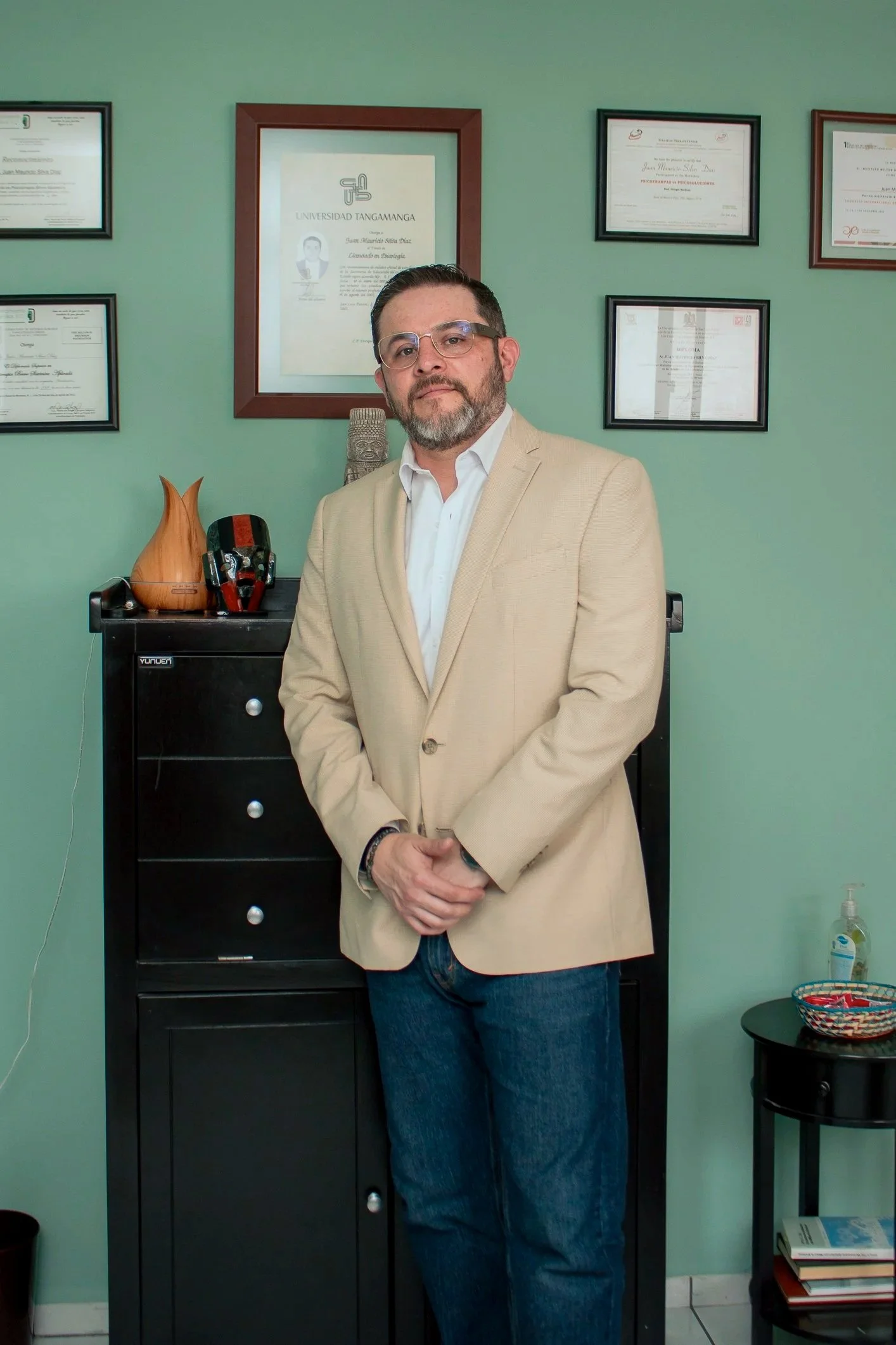 Psicologo Hombre de complexión media con barba, gafas y cabello oscuro, vestido con saco beige y camisa blanca, posando en una oficina con diplomas y certificados en la pared.