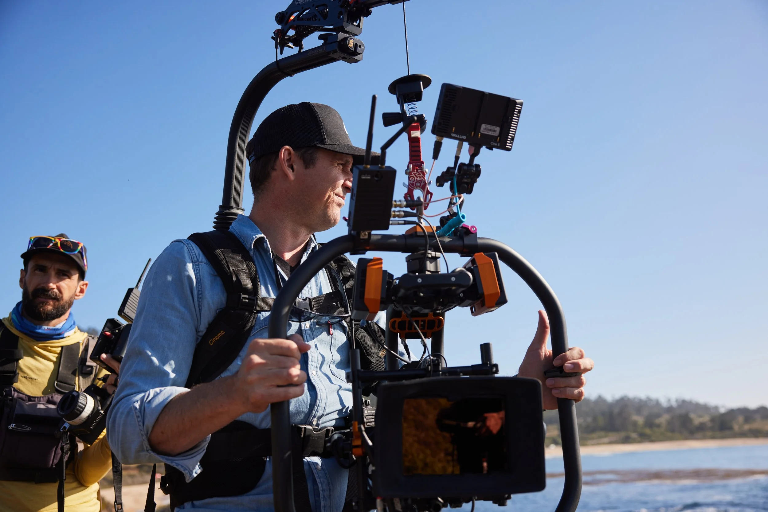 Two men working outdoors with a camera stabilizer, on a sunny day near a body of water with a shoreline and trees in the background.
