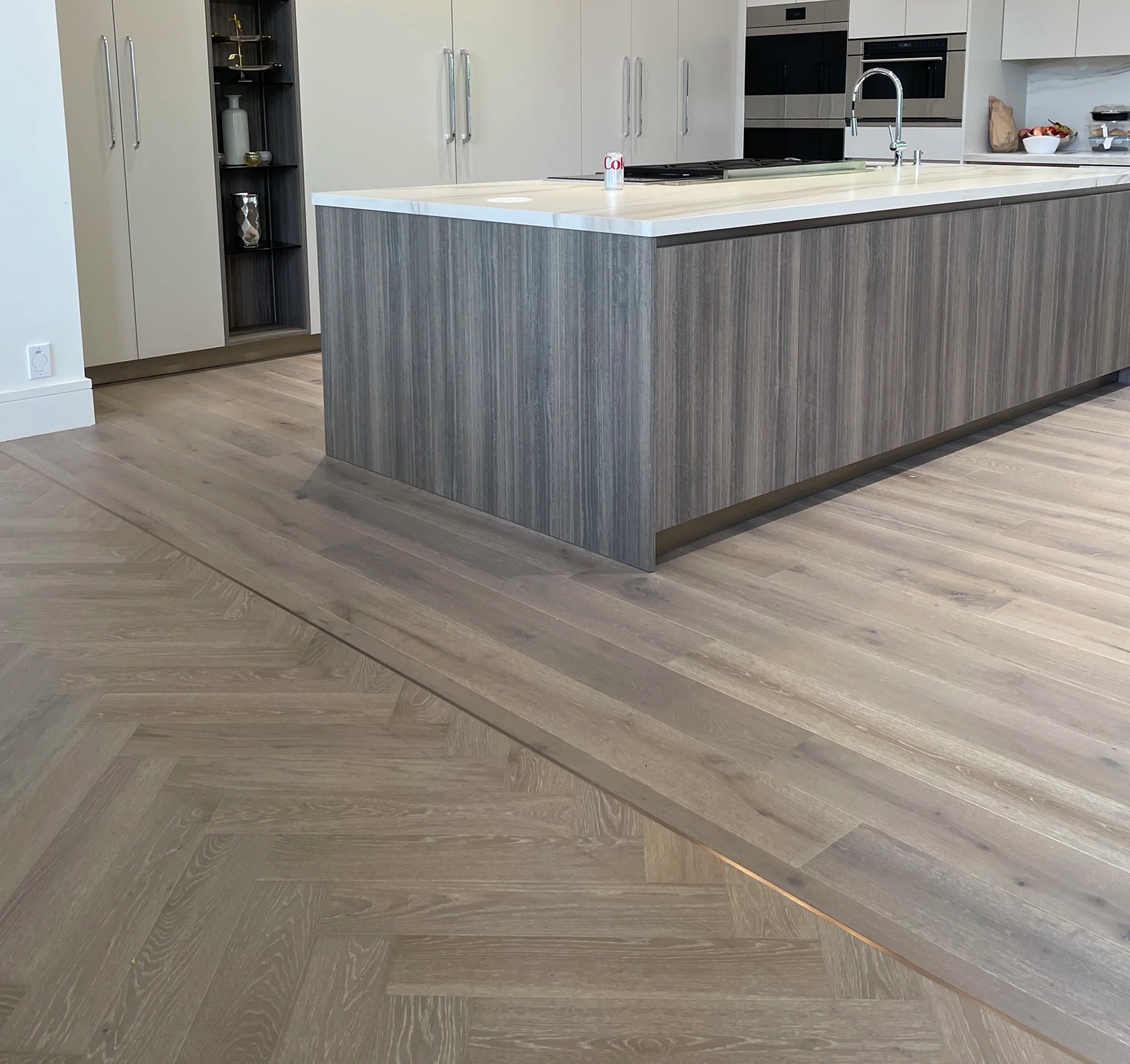 Modern kitchen interior featuring an island with a white countertop and a wood veneer front, stainless steel appliances, and light-colored cabinetry. The kitchen has light wood flooring and decorative items on the shelves and counters.