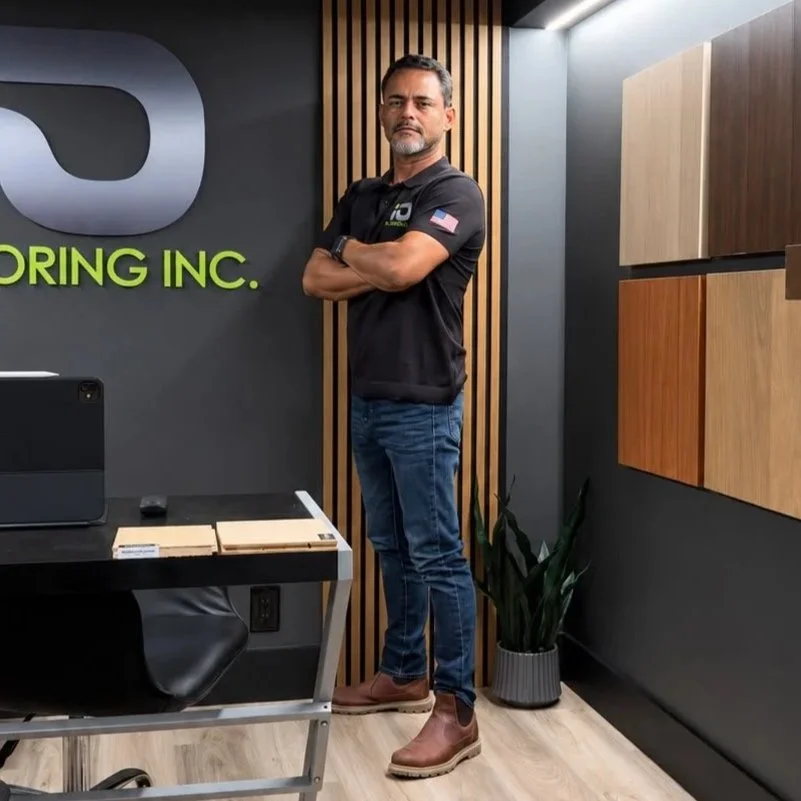 Man standing with arms crossed in an office space, wearing a black polo shirt with an American flag patch, jeans, and brown work boots, next to a wall with wood paneling and cabinets, under a sign reading ' Flooring Inc.'.
