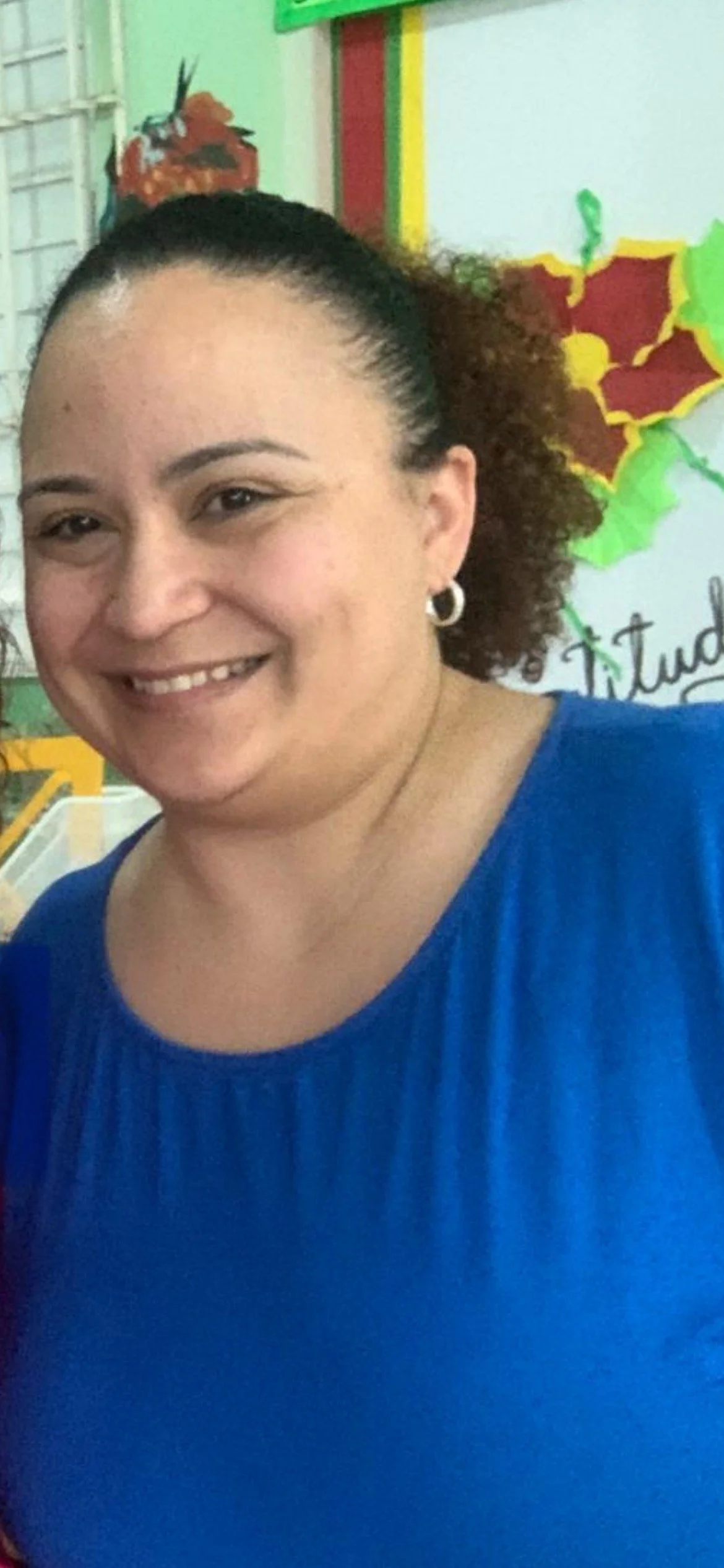 Close-up of a woman with curly hair pulled back, wearing a blue top and hoop earrings, smiling in front of colorful classroom decorations, including a poinsettia and a whiteboard.