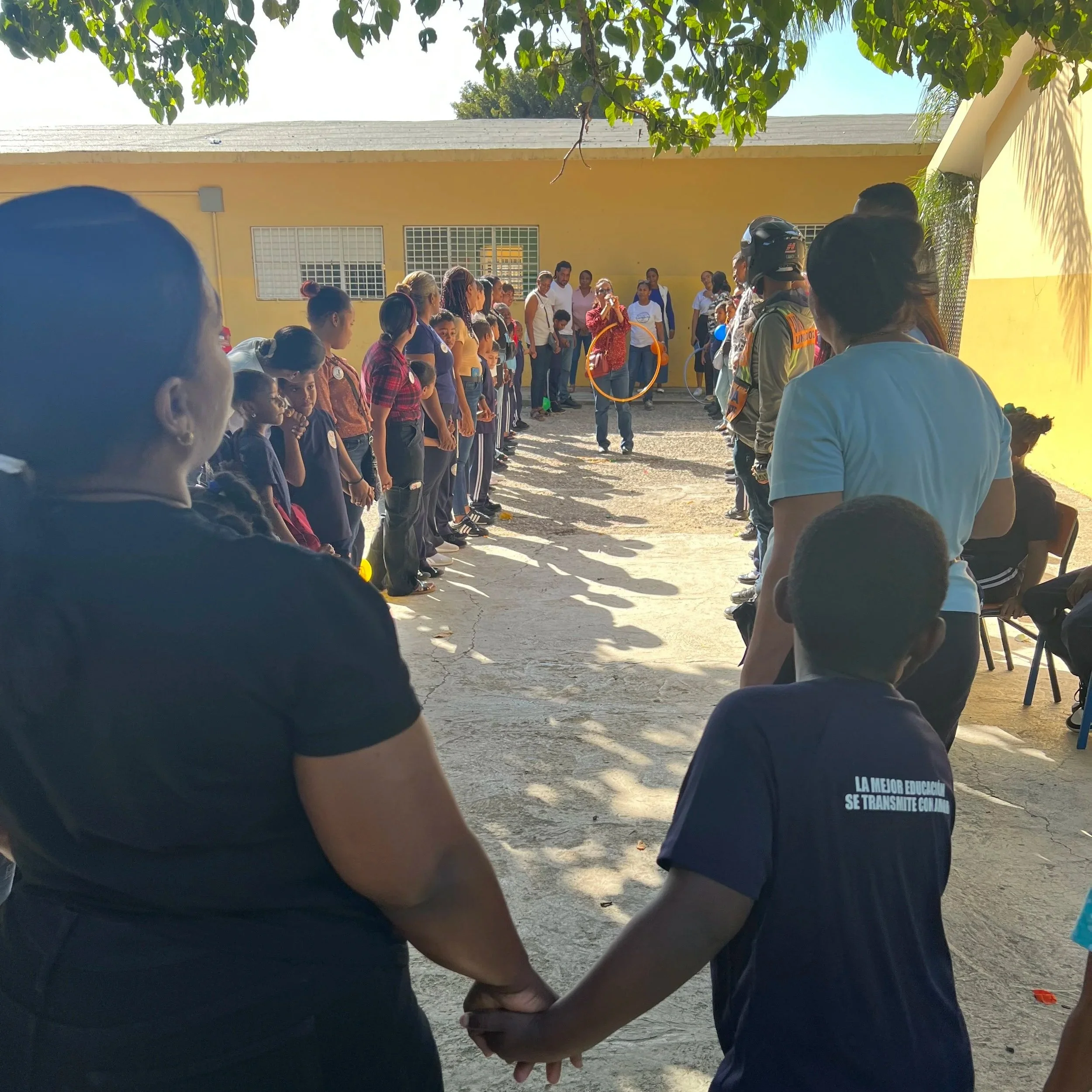 People holding hands, standing in a circle outdoors, with a yellow building in the background, several children, and adults participating in a community event or gathering.
