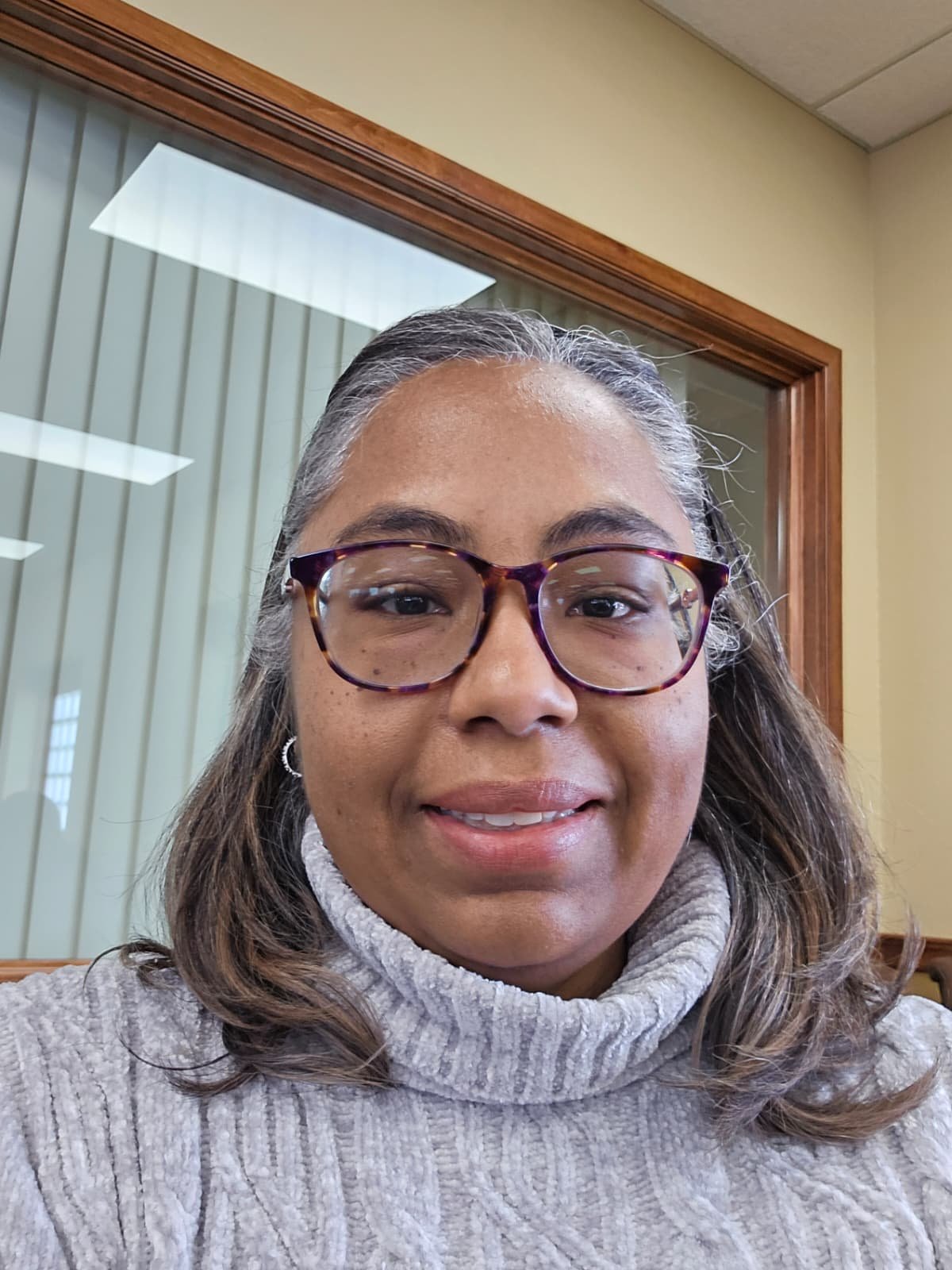 A woman with long, wavy hair and glasses taking a selfie in an office setting with a window and vertical blinds in the background.