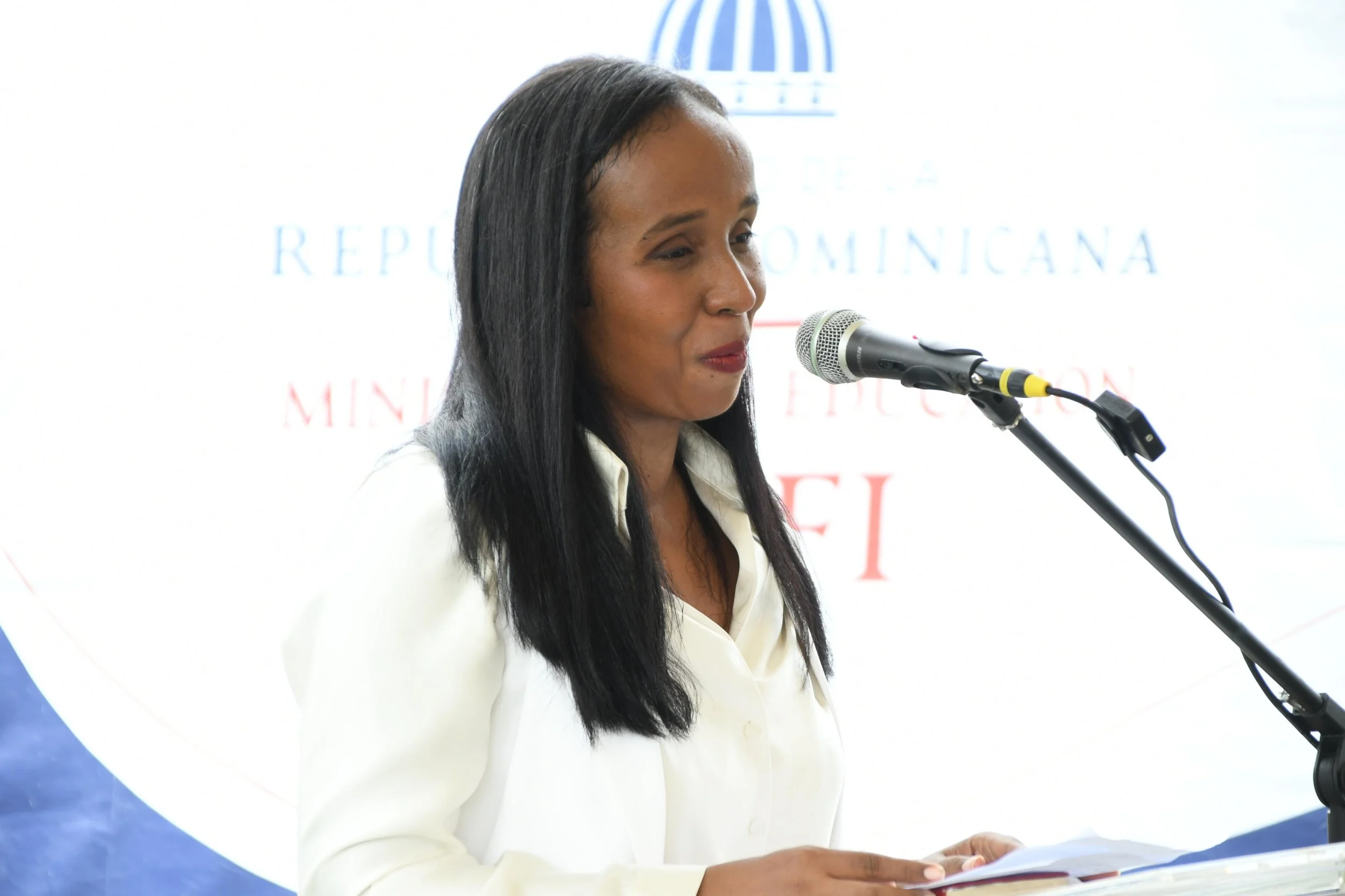 A woman with long black hair in a white blazer speaking into a microphone at a presentation or press event, with a blurred logo and text in the background.