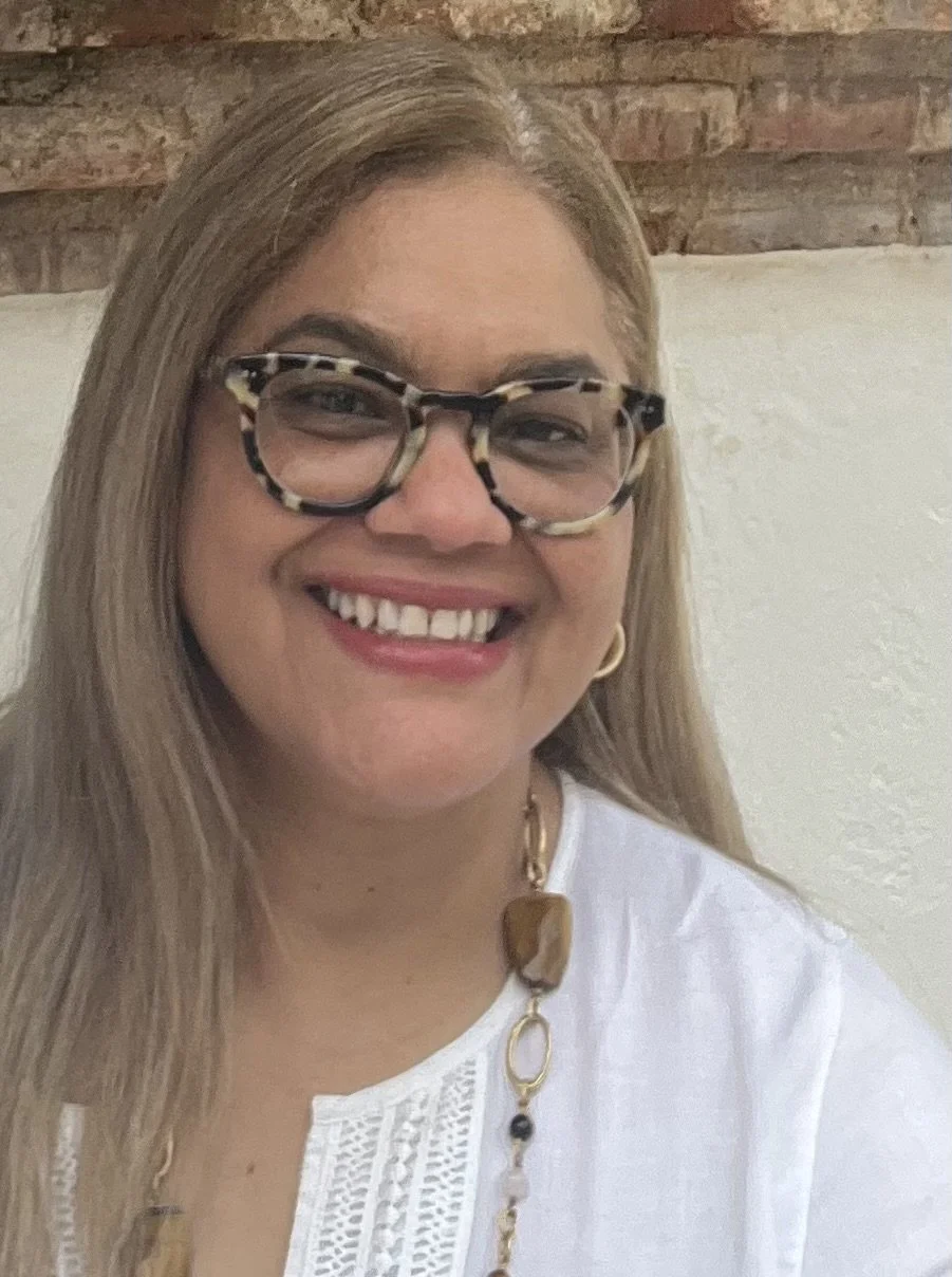 A woman with long blonde hair, wearing glasses with a tortoiseshell pattern, smiling, and wearing a white top with a gold necklace that has large beads.